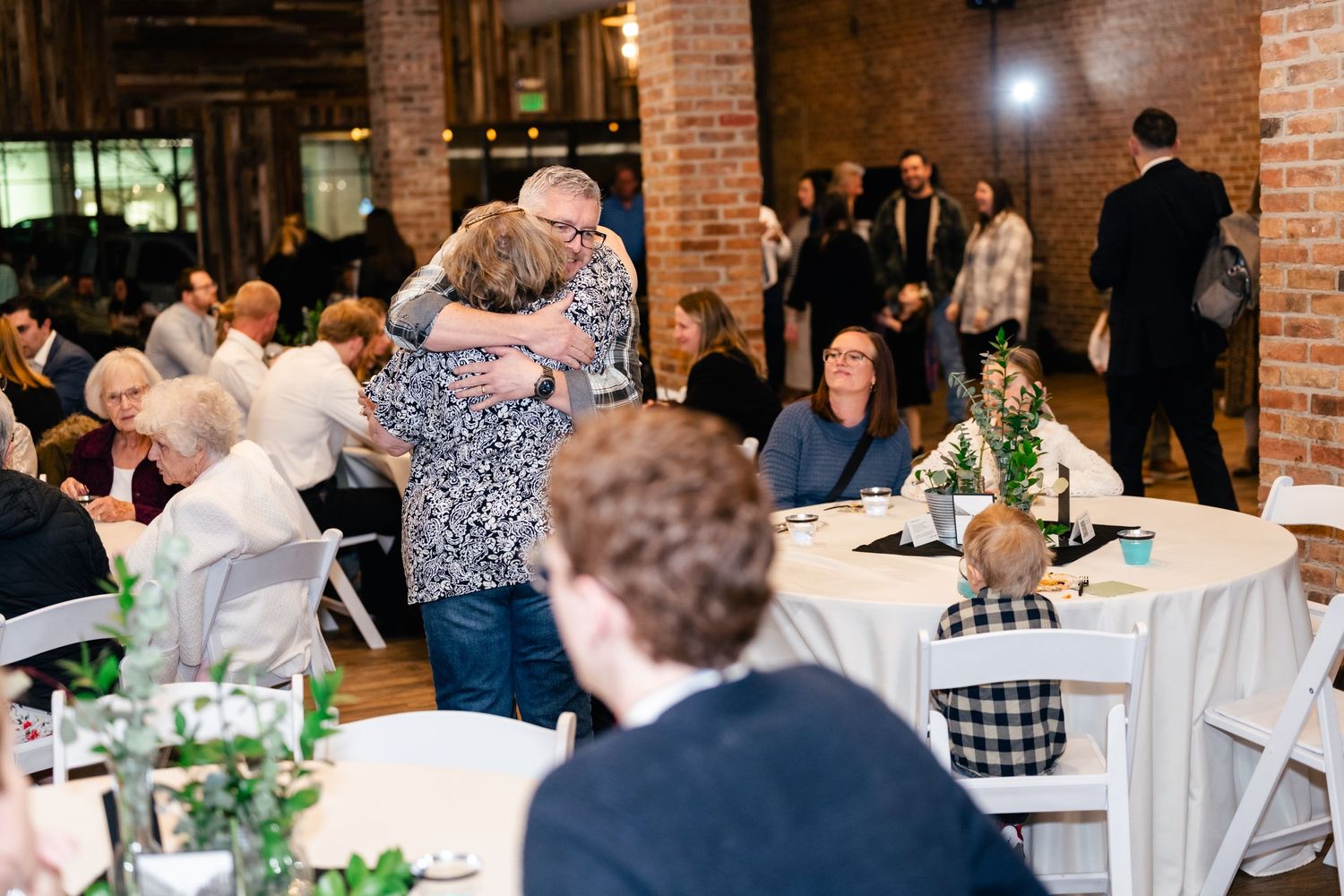 Wedding reception guests share embraces inside a rustic brick-walled venue with round tables and greenery centerpieces.