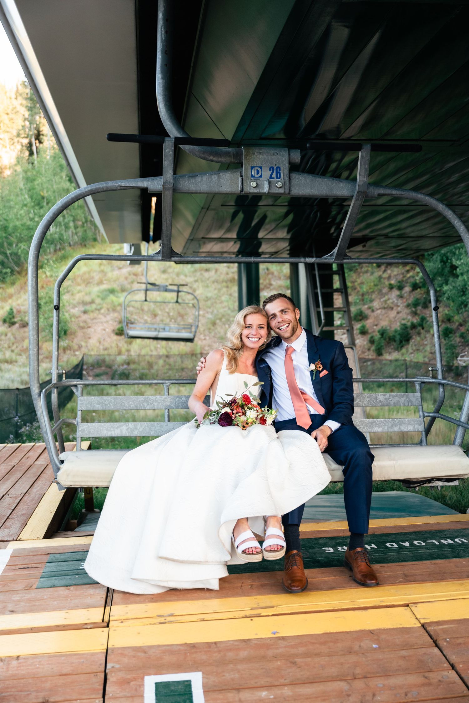 Happy newlyweds sit together on ski lift chair with bride's dress flowing and mountain scenery behind. Park City Wedding Photographer.