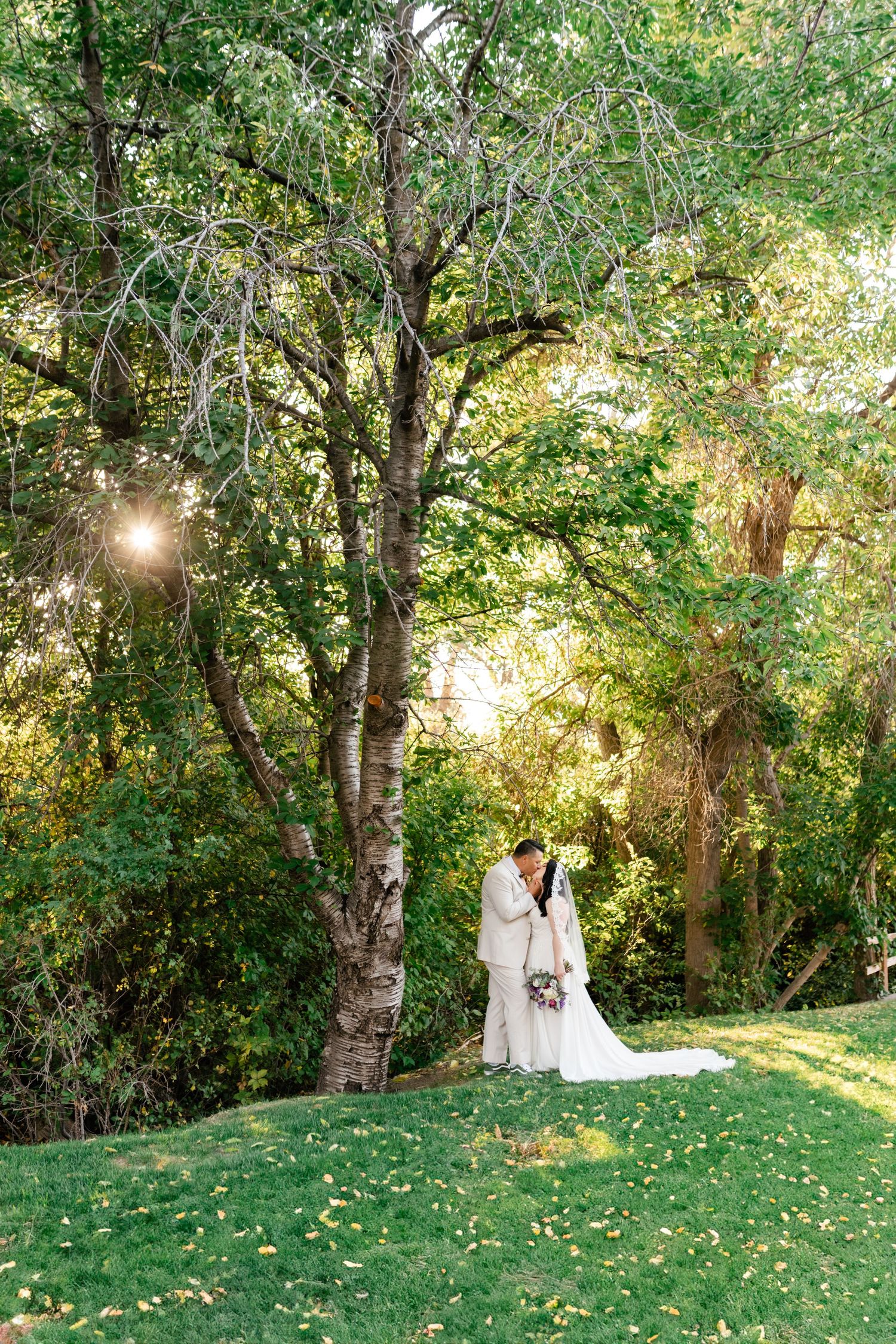 Couple embraces under sunlit trees in a lush garden setting during an outdoor wedding ceremony.