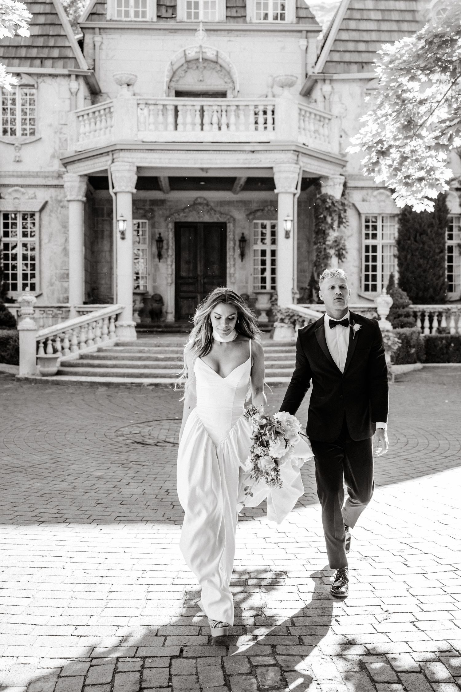 Black and white photo of wedding couple exiting luxurious mansion with grand pillared entrance and ornate balcony.