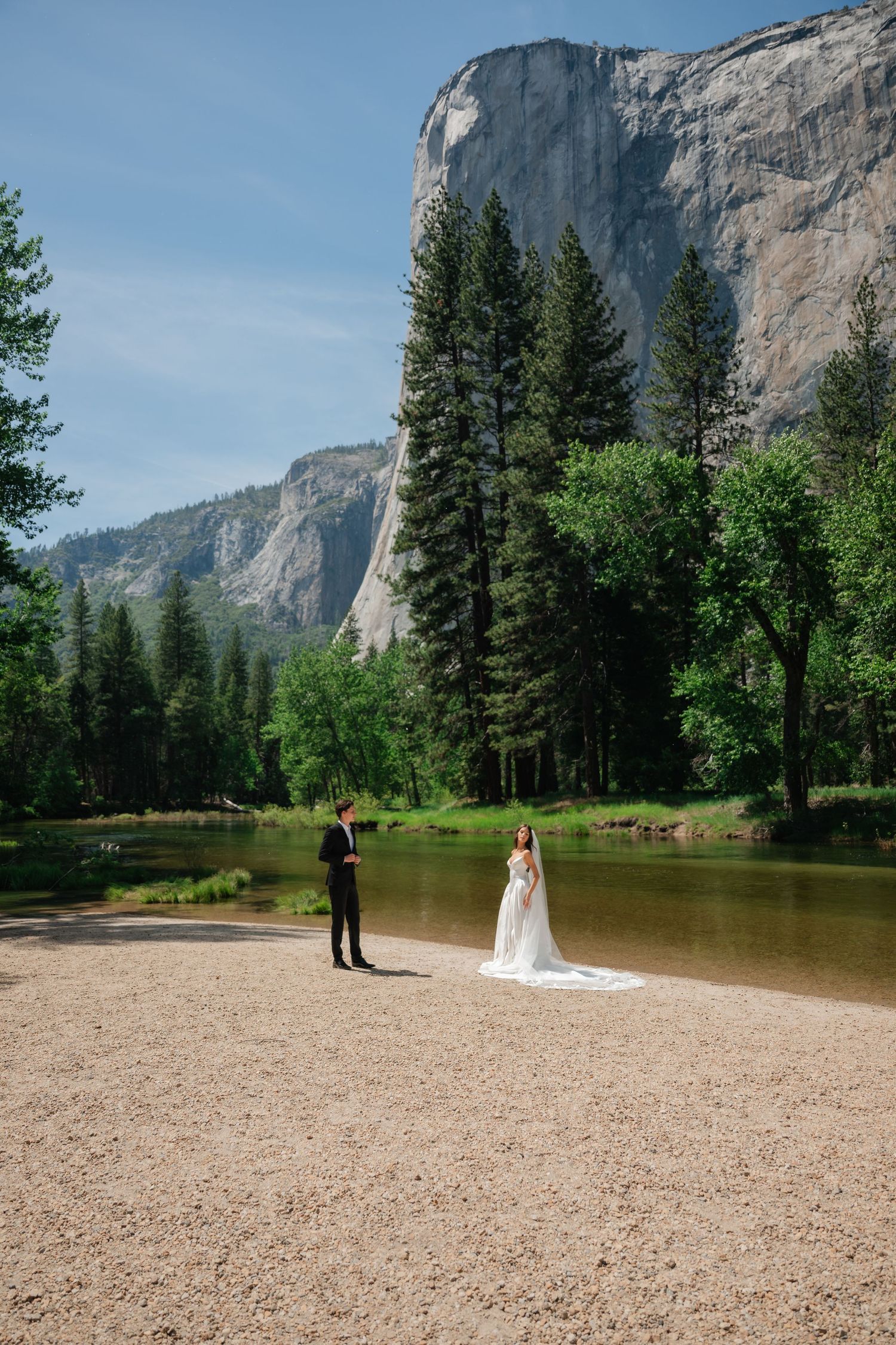 Bride and groom stand apart on sandy shore at Yosemite with El Capitan and pine trees backdrop.