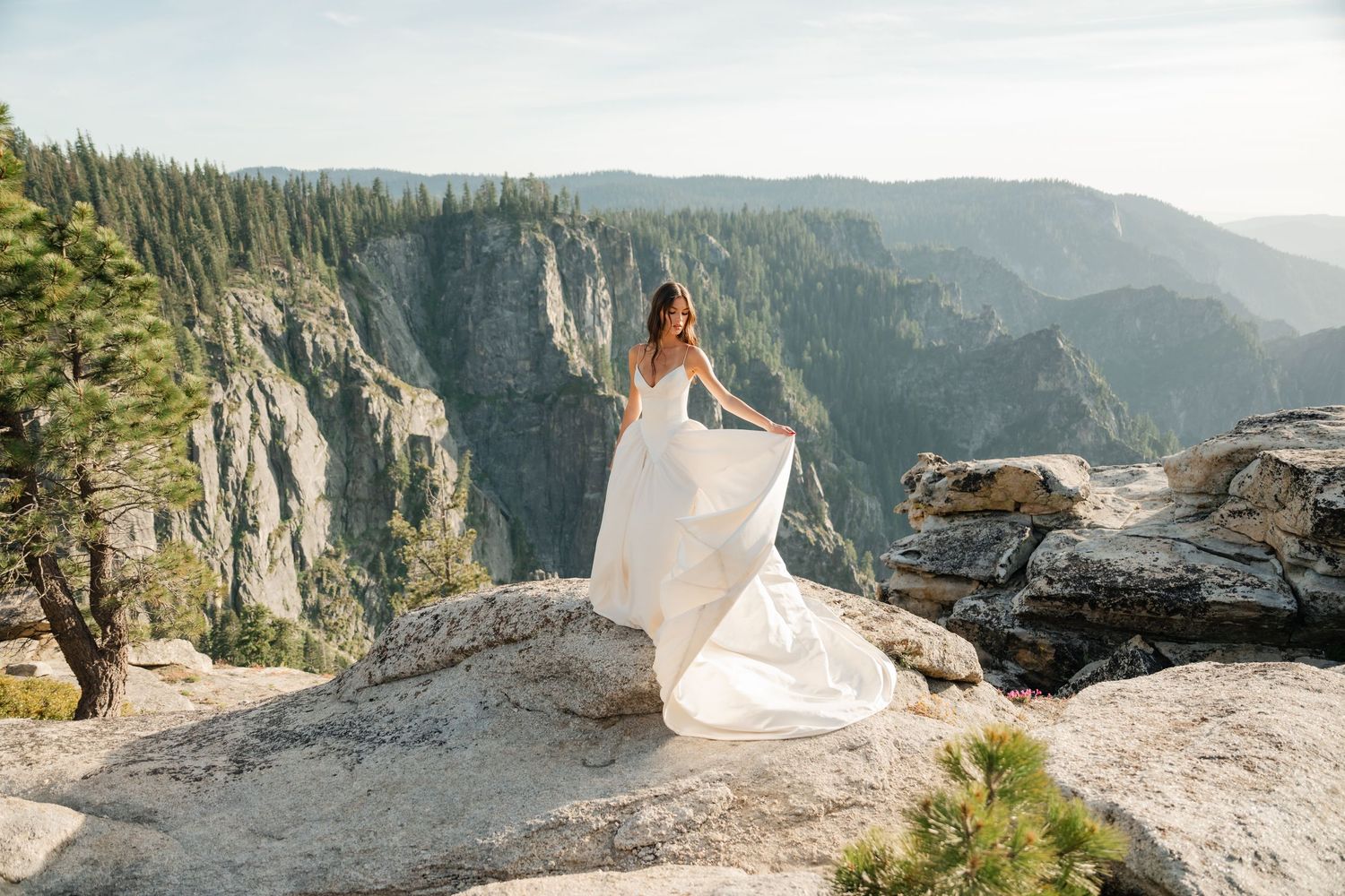 Bride in flowing white gown poses on rocky cliff at Yosemite with stunning mountain valley views.