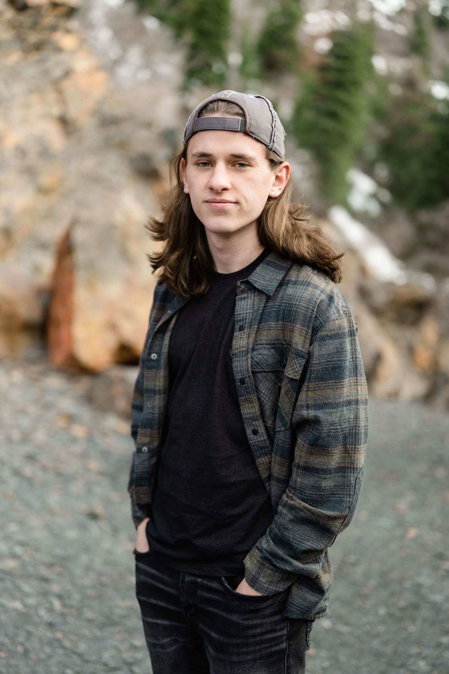 Person in a plaid flannel jacket and baseball cap poses casually against a rocky outdoor backdrop.