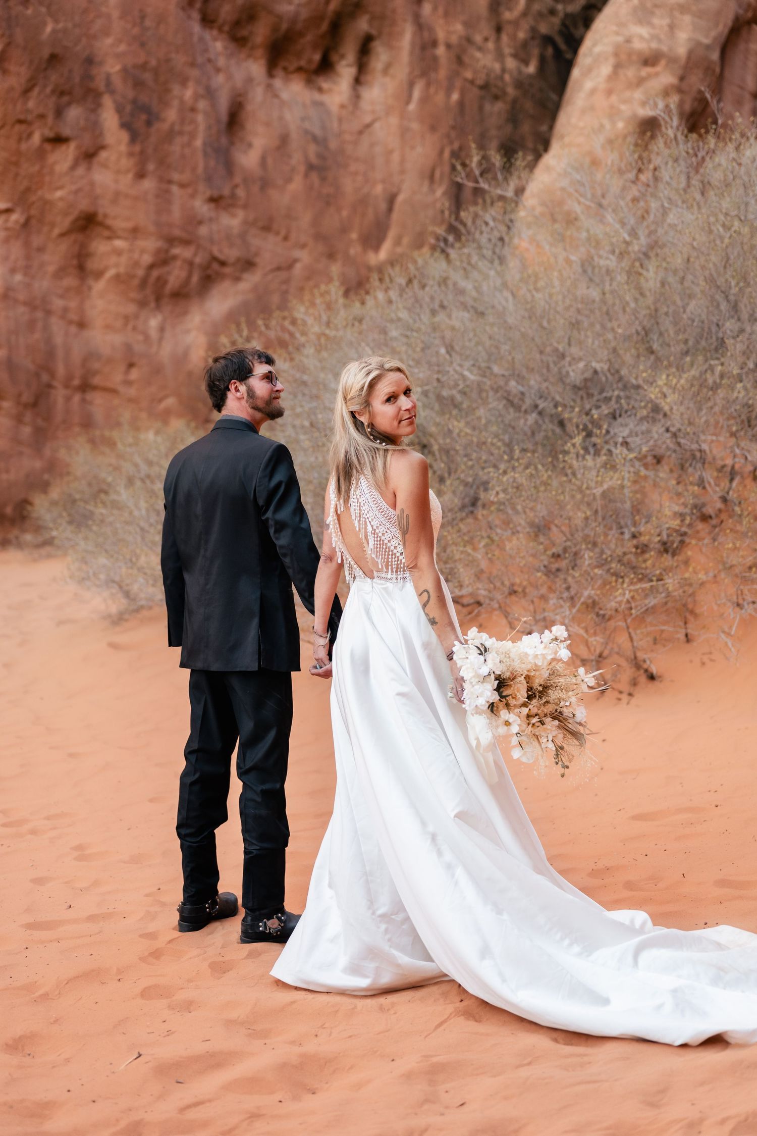 A wedding couple poses against red rock formations in a desert landscape with flowing white dress and black suit.