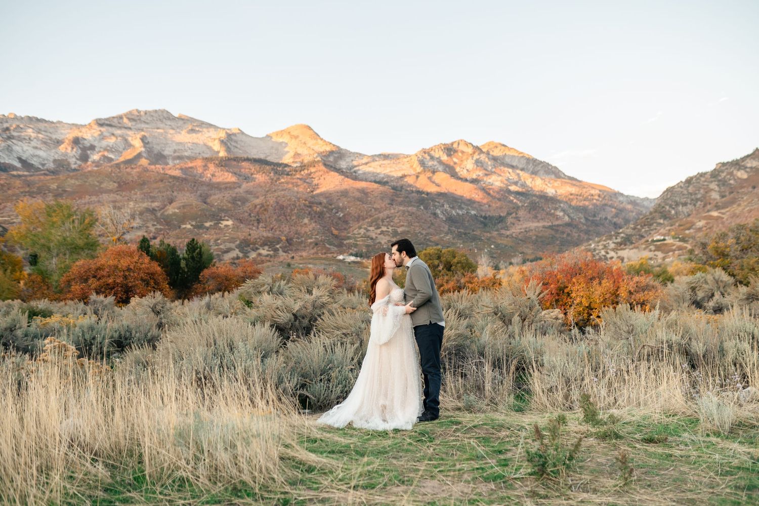 Couple embraces in front of scenic mountain landscape with autumn foliage and desert brush at sunset.