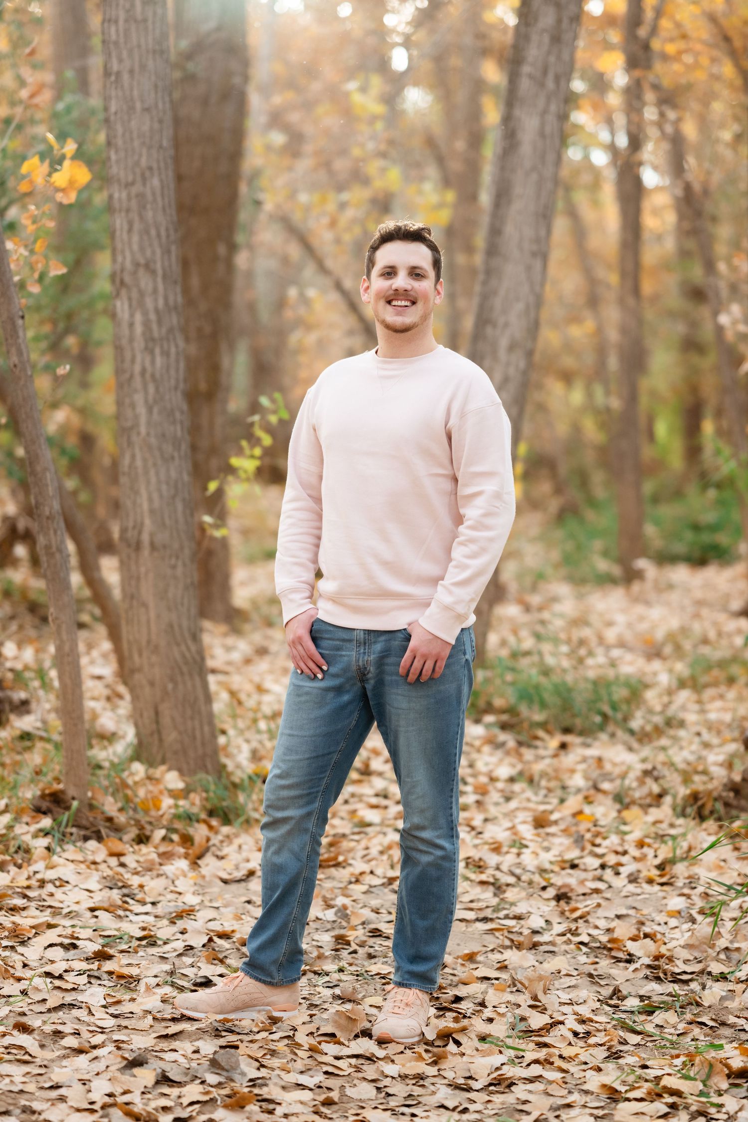 Person in light colored sweater and jeans poses playfully among fallen leaves in autumn forest.