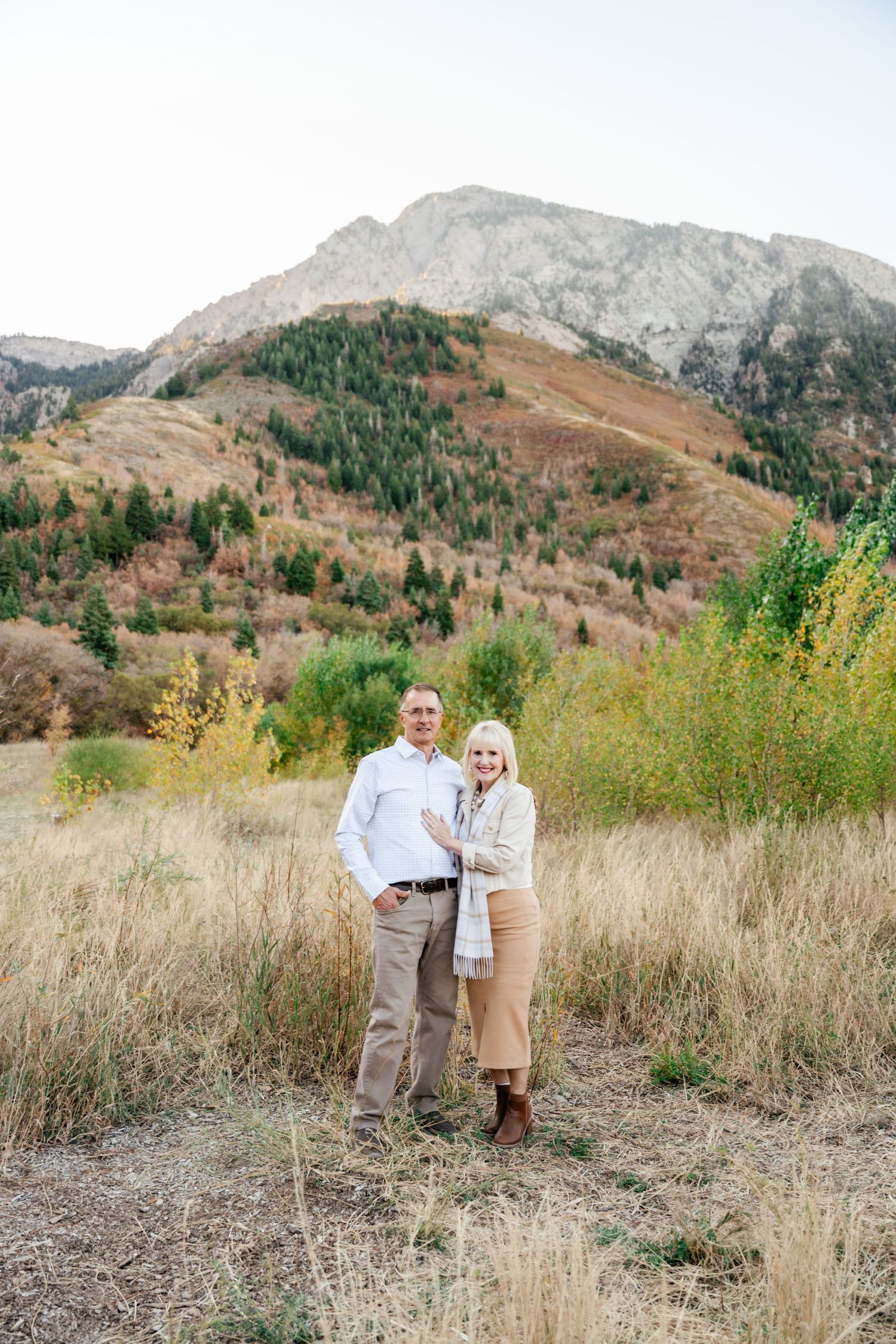 Couple standing together in a scenic mountain meadow with autumn foliage and rocky peaks in the background.