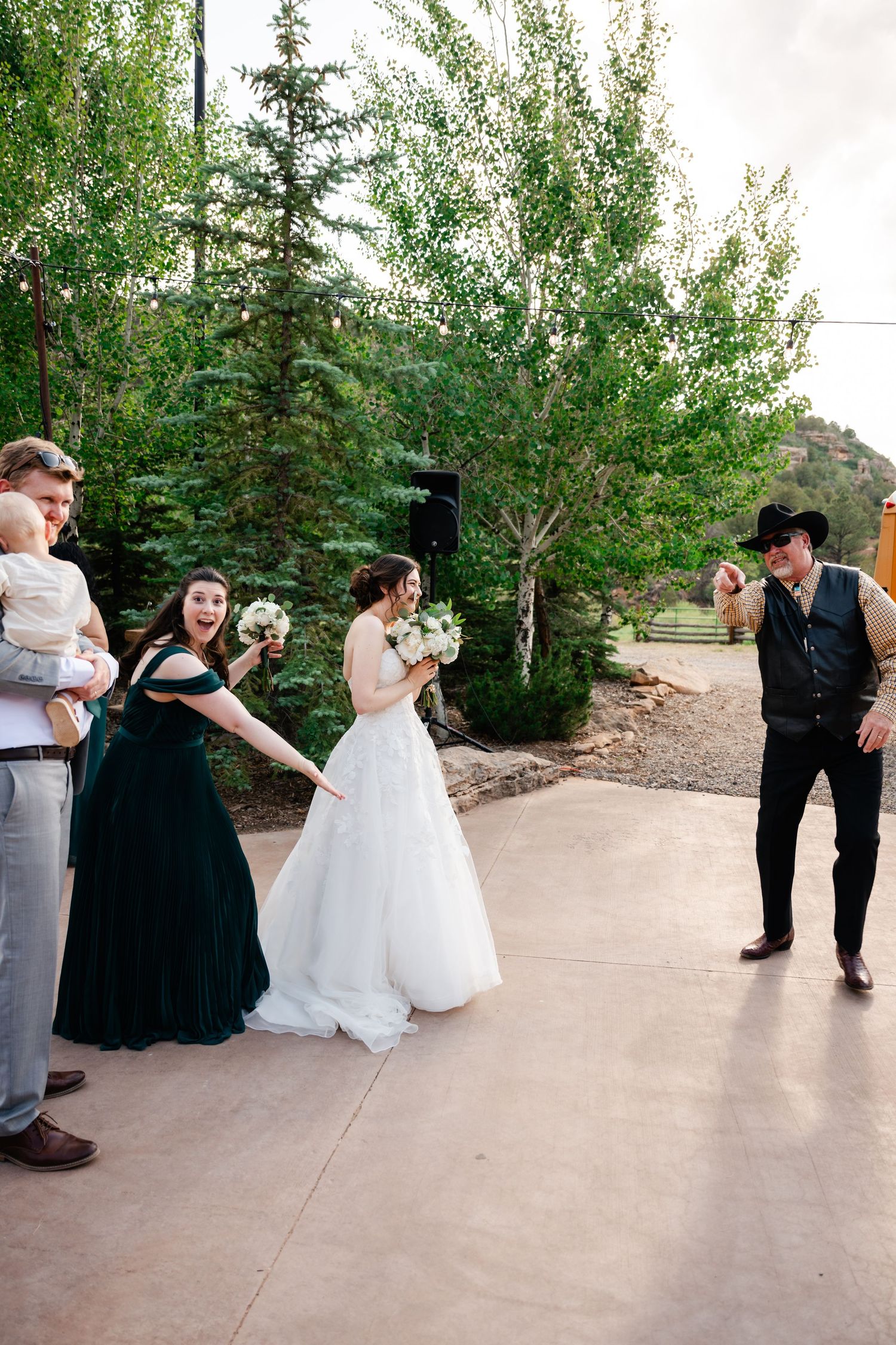 Wedding party members dancing in formal attire on an outdoor patio surrounded by greenery.