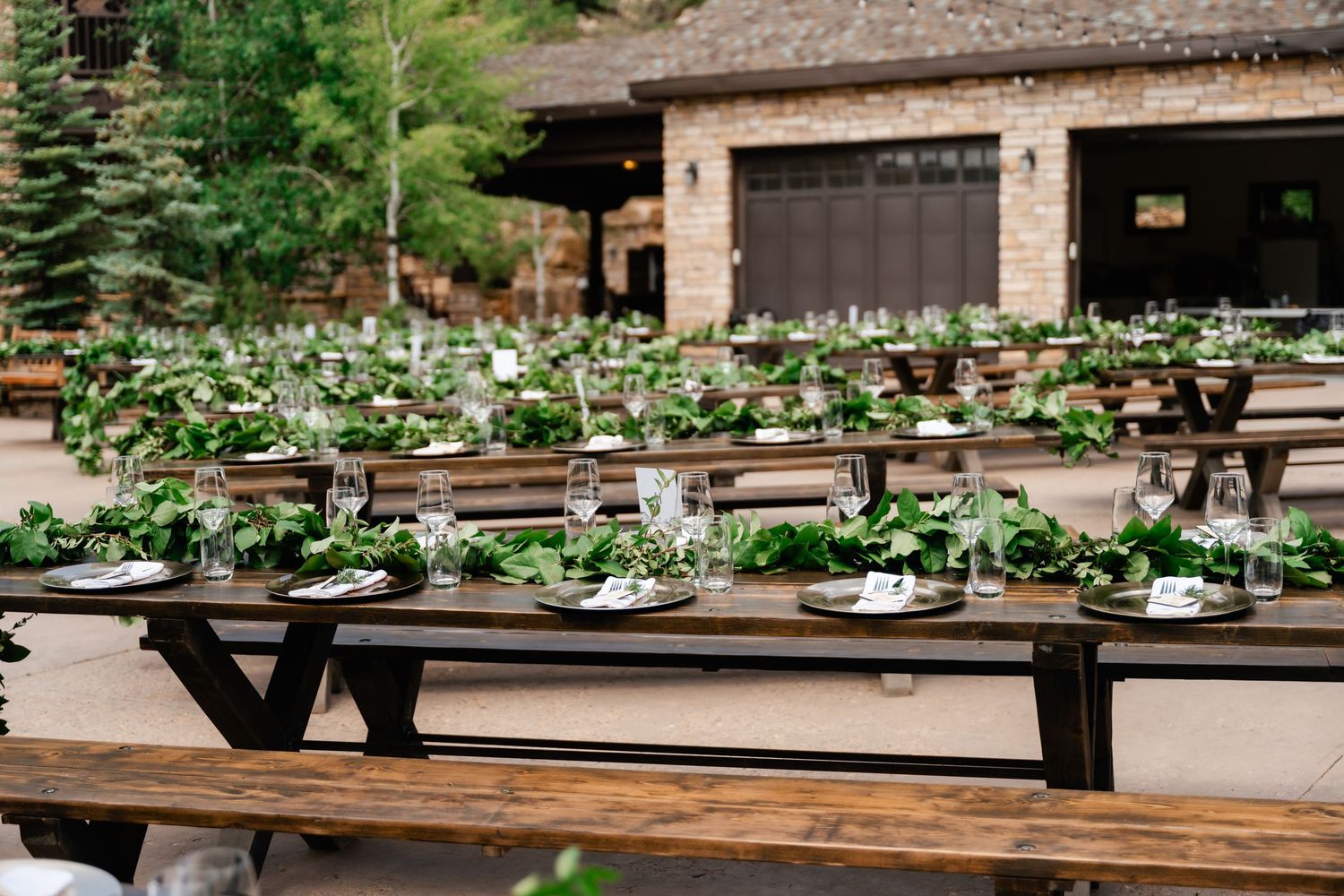 Rustic outdoor wedding reception setup with long wooden tables and benches surrounded by lush greenery.