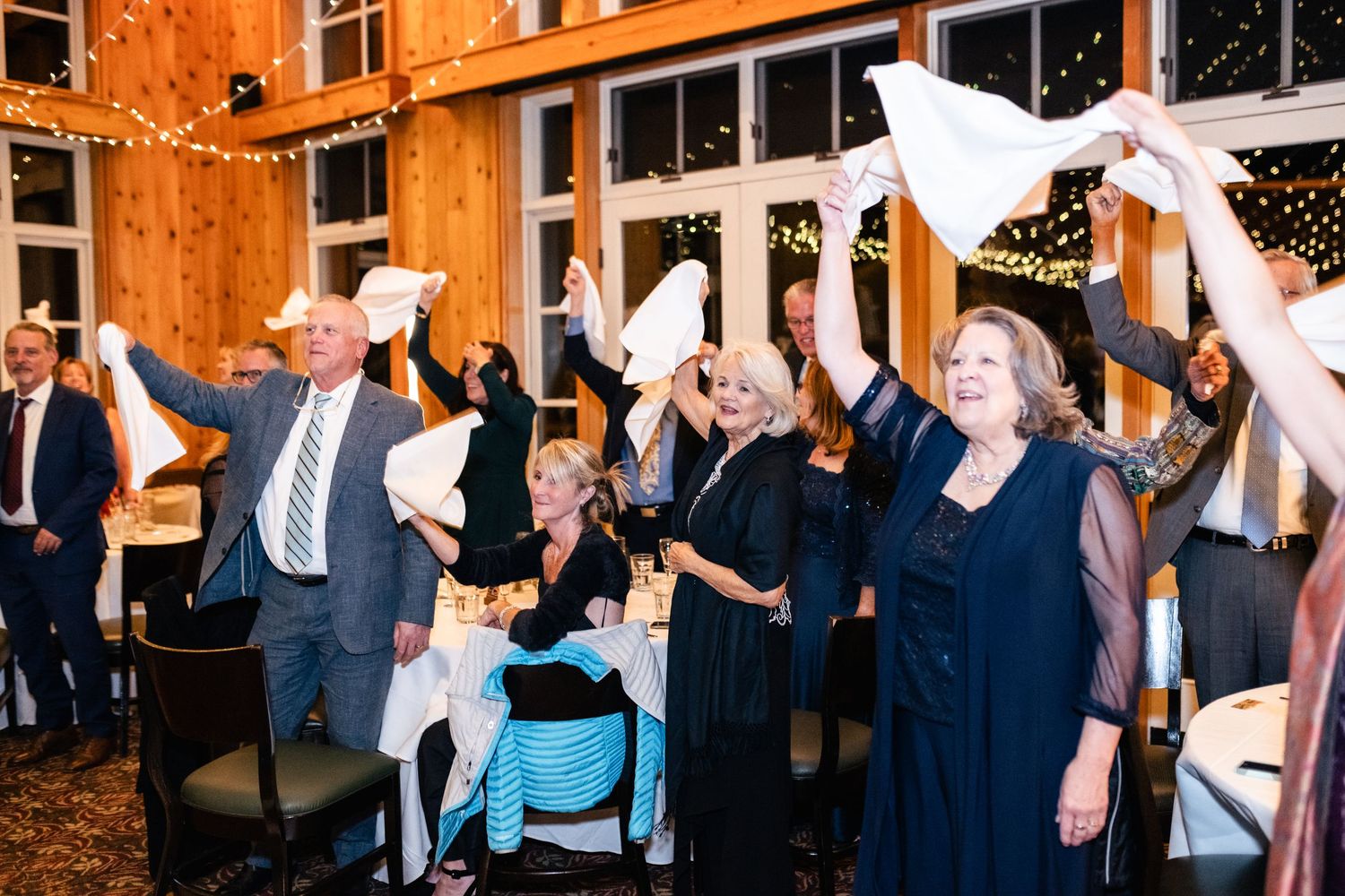 Wedding reception guests waving white napkins in rustic venue with string lights.