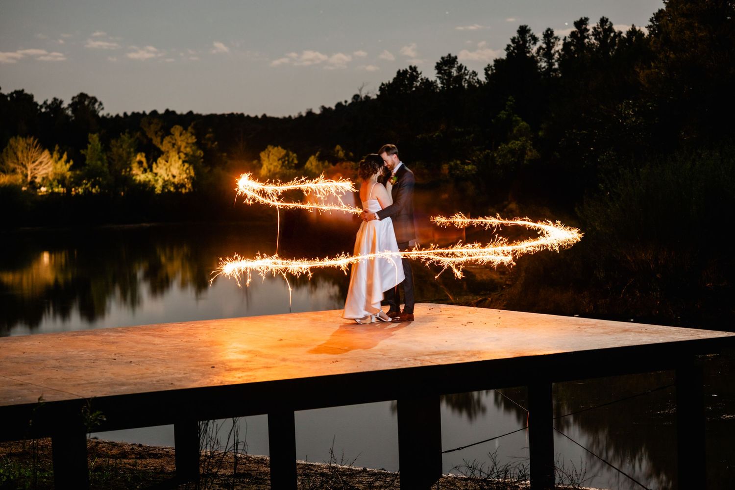A couple dances on a dock at dusk surrounded by glowing string lights reflecting on the lake.