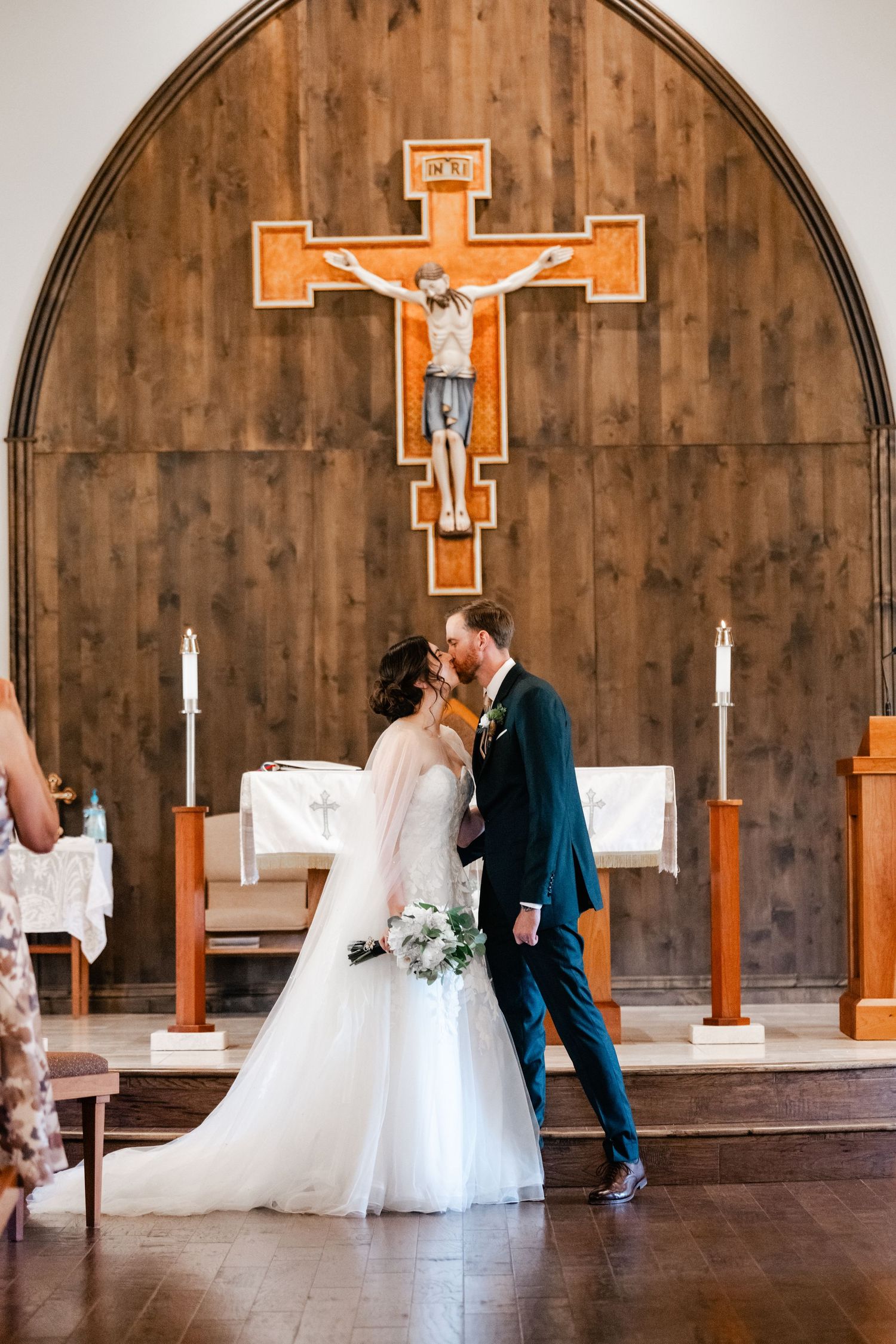 A couple shares their first kiss at the altar beneath a wooden crucifix in a rustic church setting.