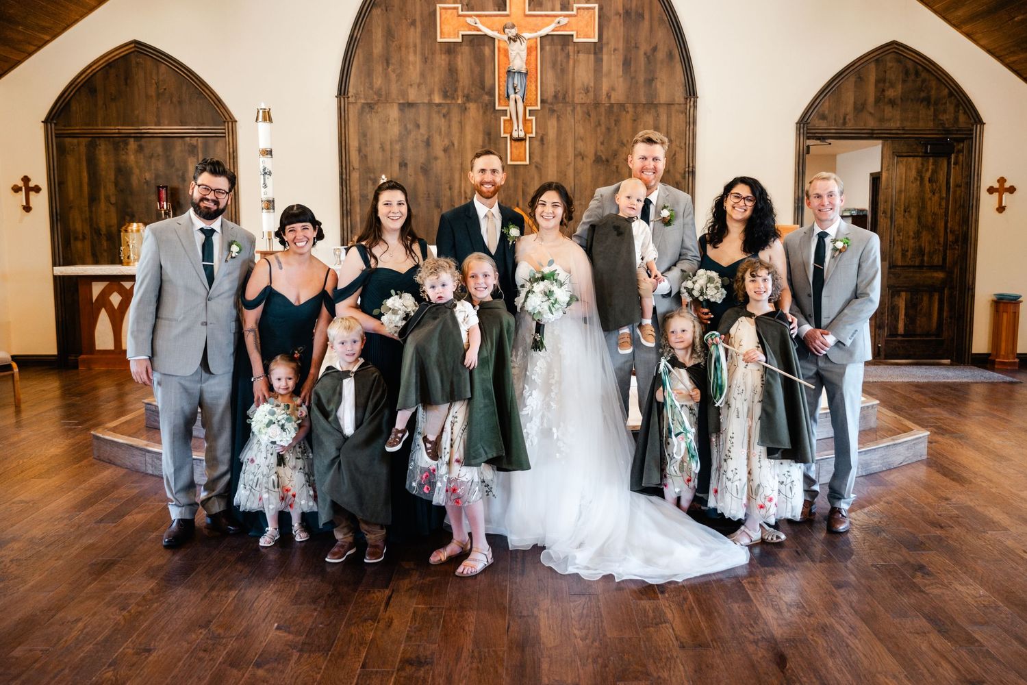 A wedding party poses together inside a church with wooden floors and arched doorways beneath a cross on the wall.