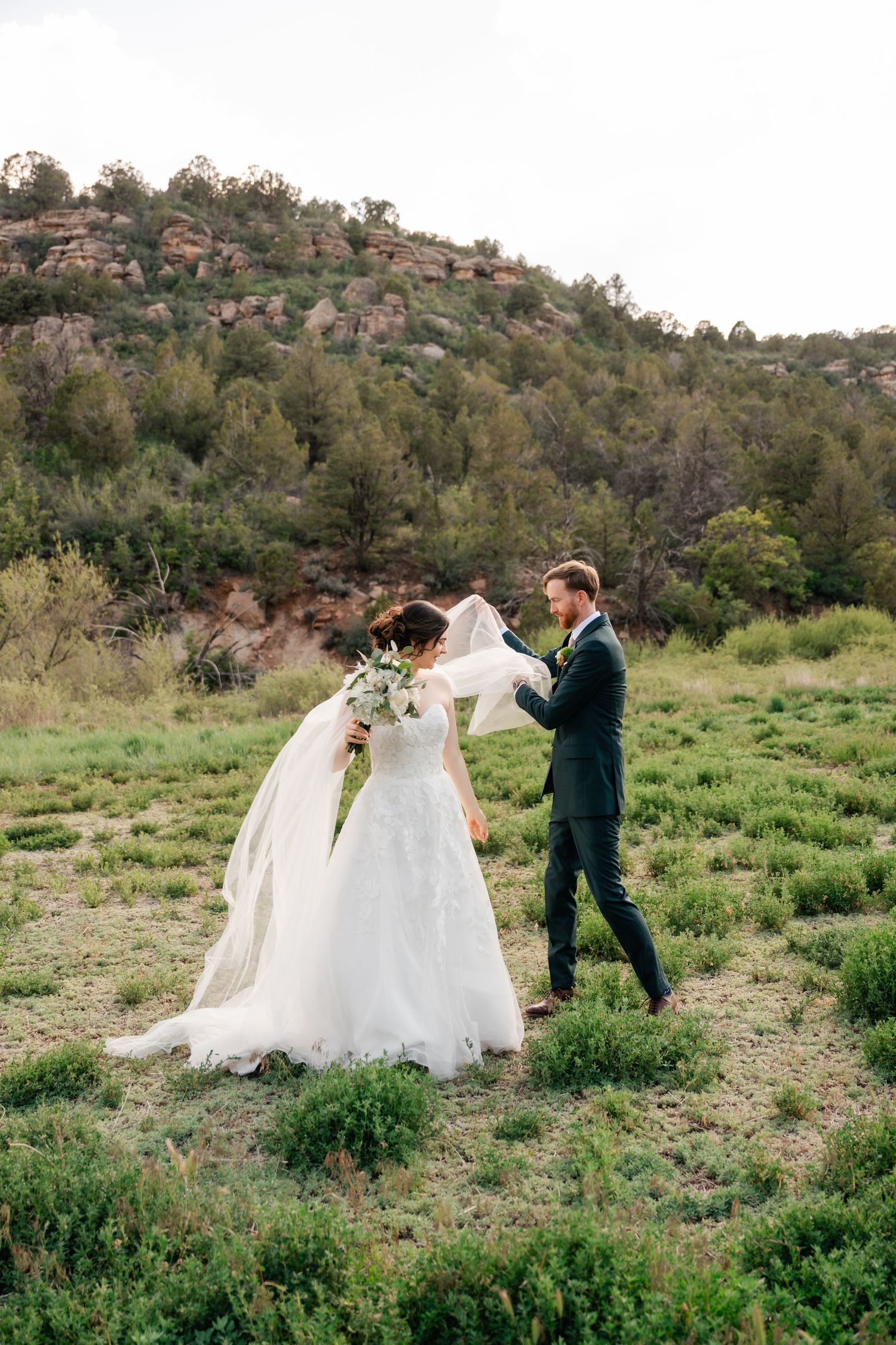 A couple in wedding attire walks through a scenic desert hillside with green brush and rocky terrain in the background.