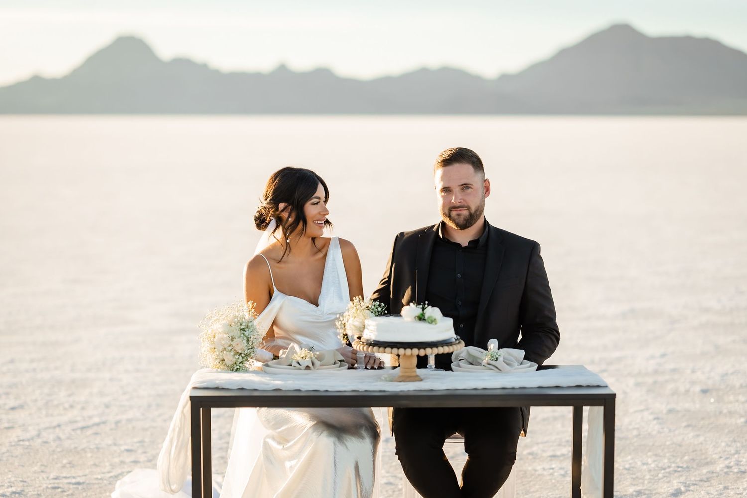 Elegant wedding reception setup on salt flats with mountain backdrop at sunset.