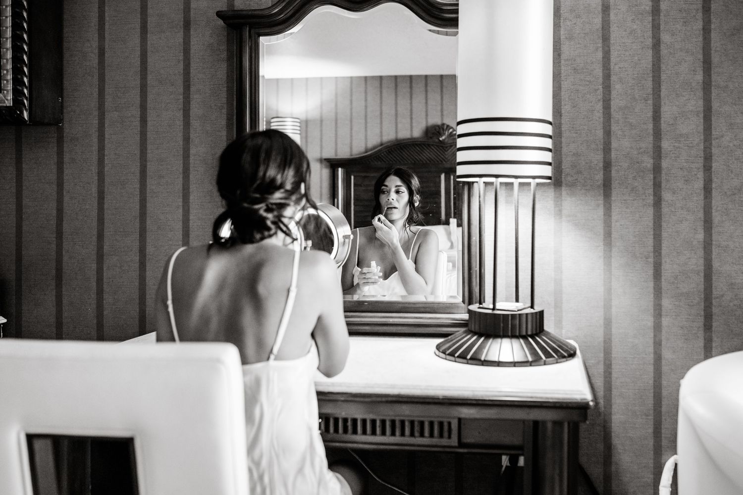A black and white photograph shows a reflection in a vintage vanity mirror with an ornate table lamp.