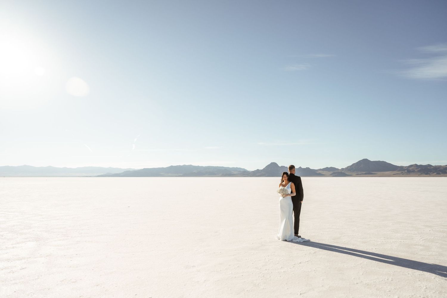 A couple stands together on the vast white expanse of the Bonneville Salt Flats in Utah during a bright sunny day.