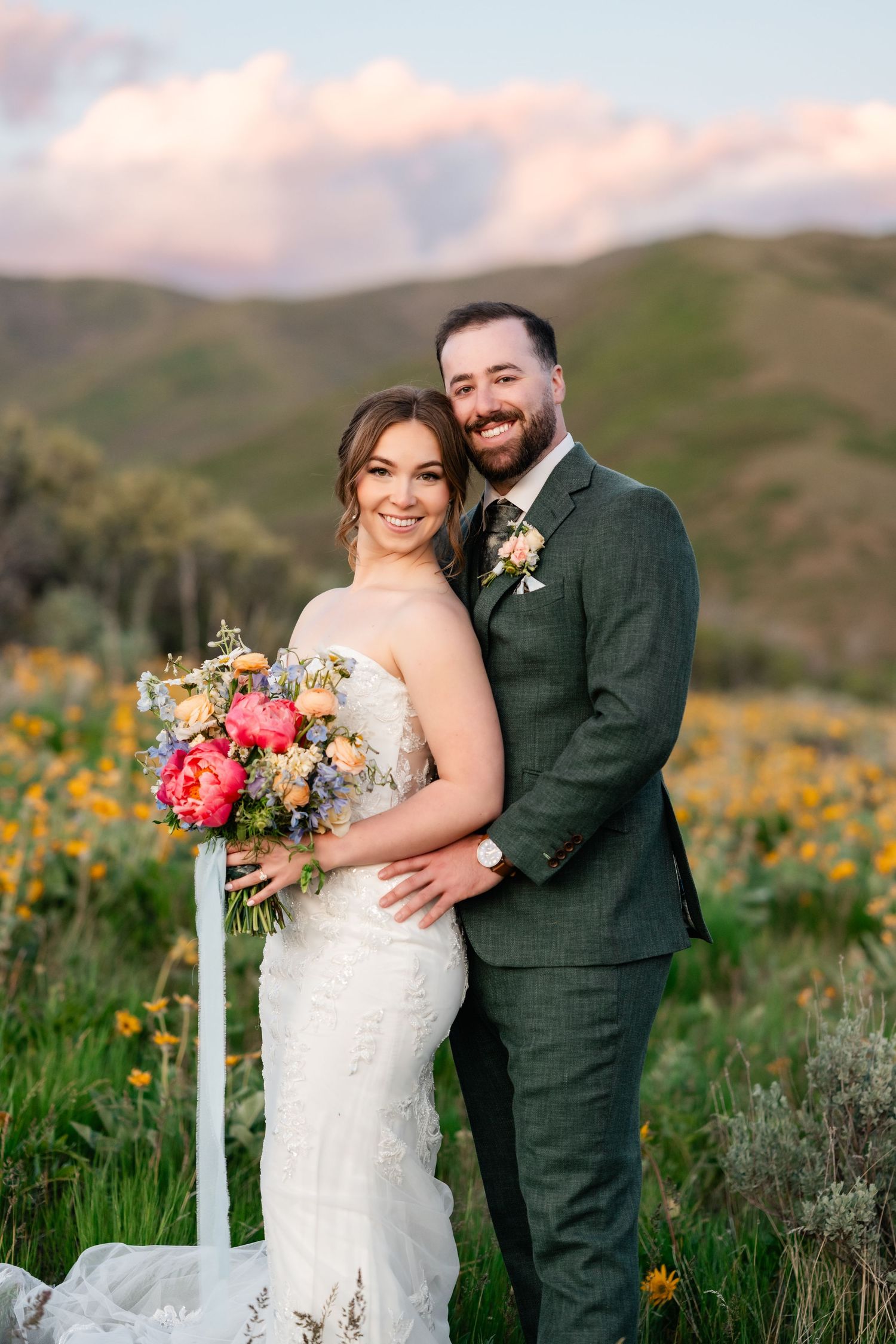 Joyful wedding couple embraces in field of spring flowers with mountains in background.