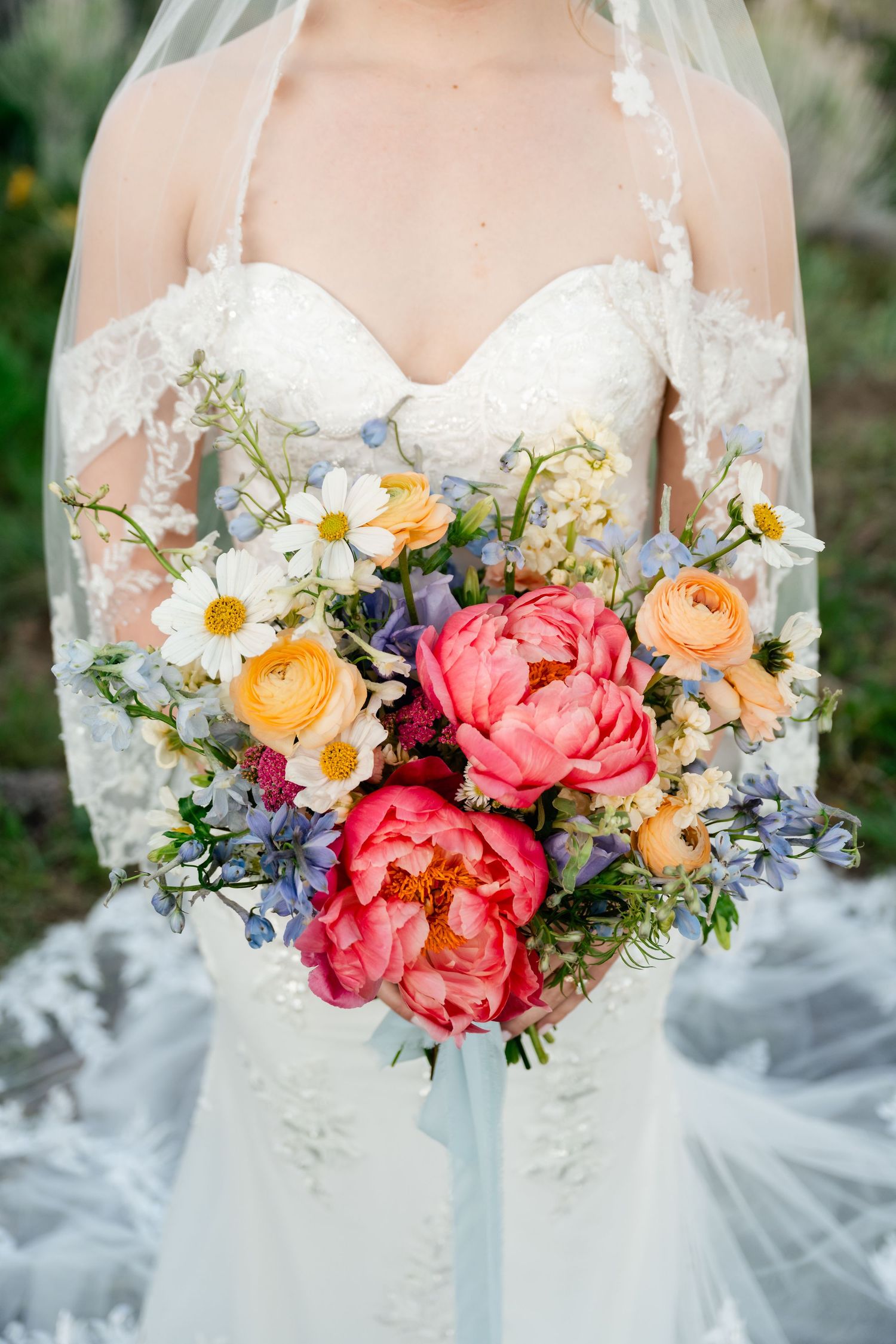 Bridal bouquet close-up with coral peonies, daisies and delicate blue flowers against white dress.