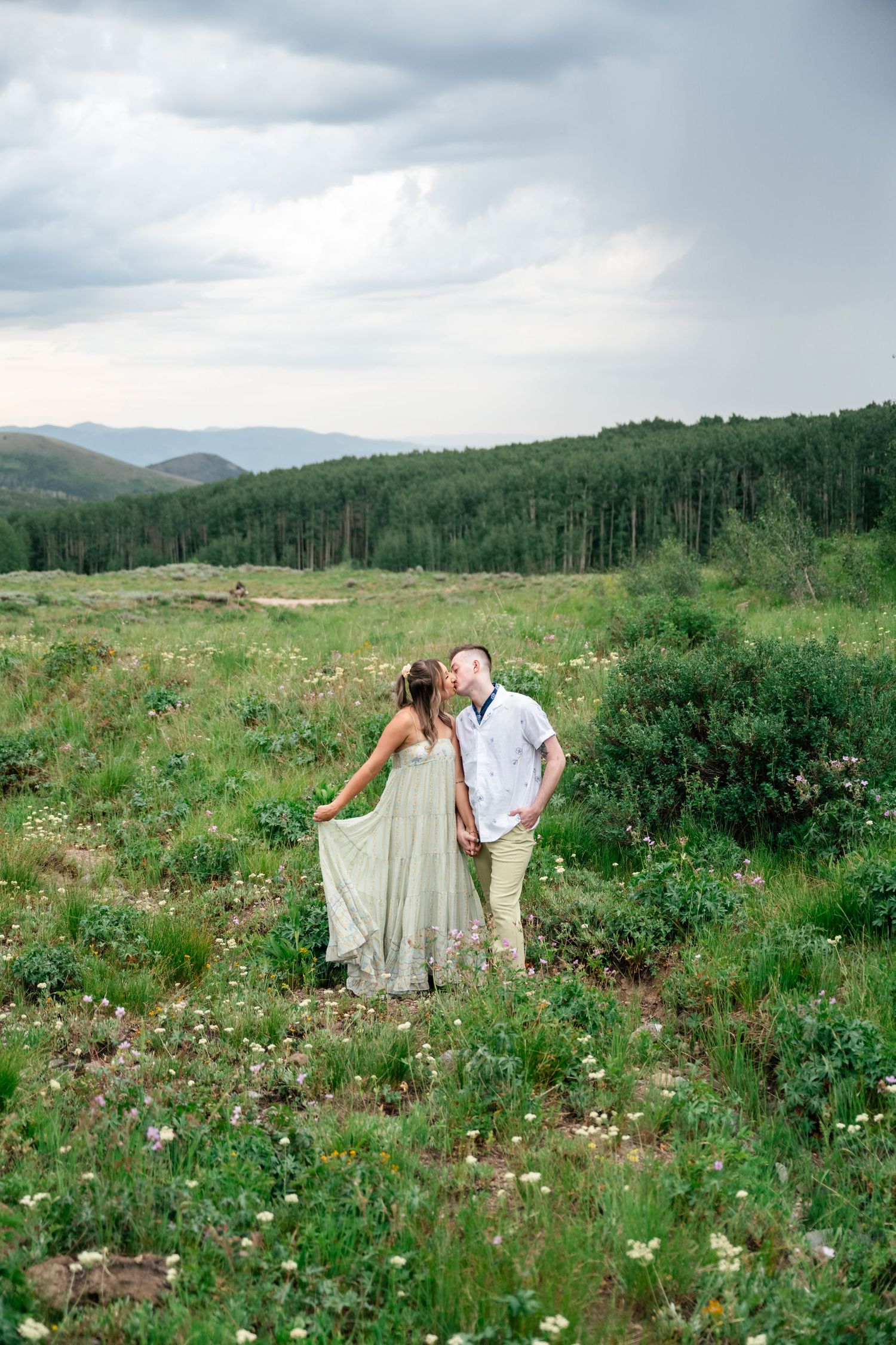 A couple stands together in a wildflower field with mountains and forests in the background.