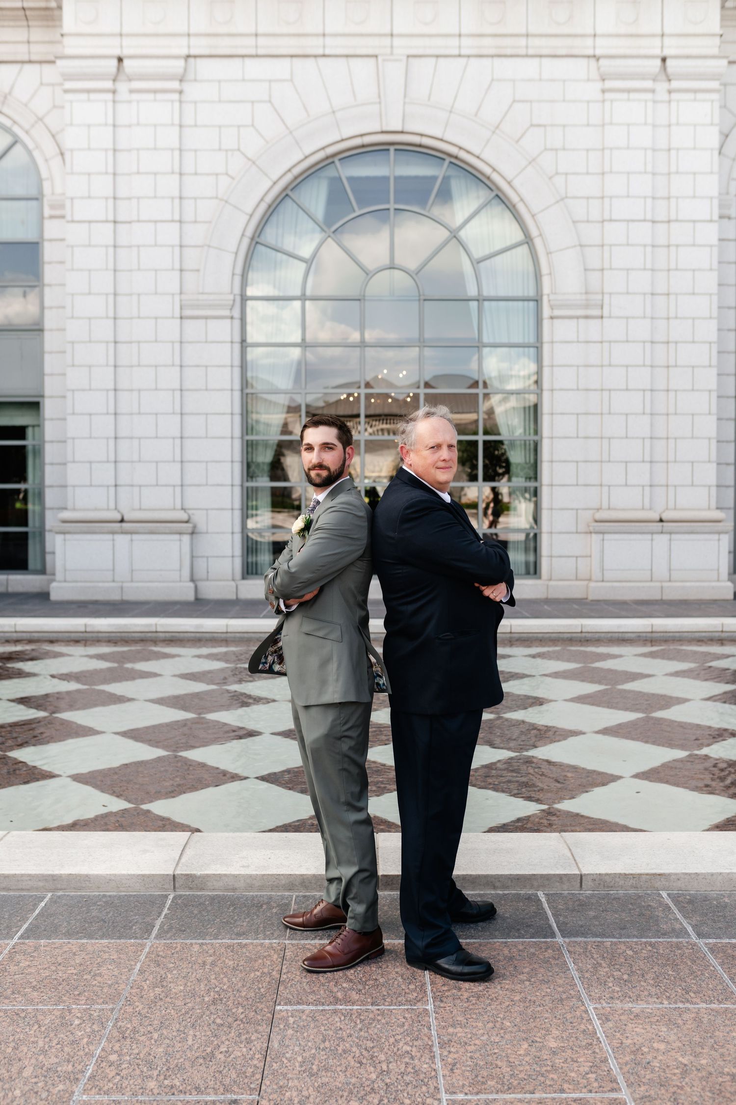 Two businessmen stand back-to-back in front of a grand arched window on a checkered marble floor.