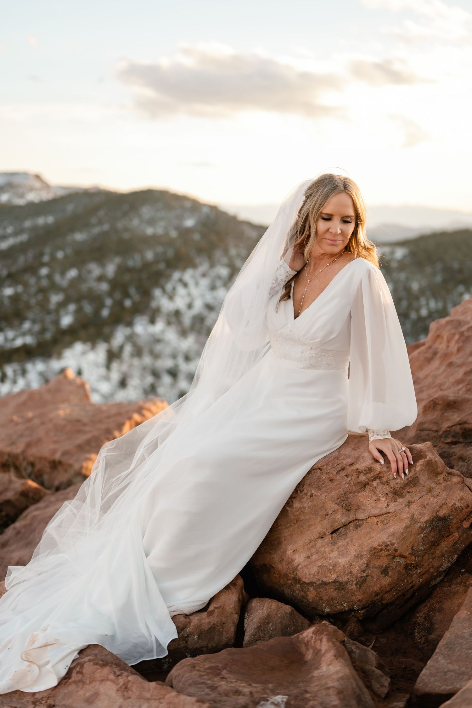 Bride in flowing white dress poses gracefully on red rock formations at sunset.