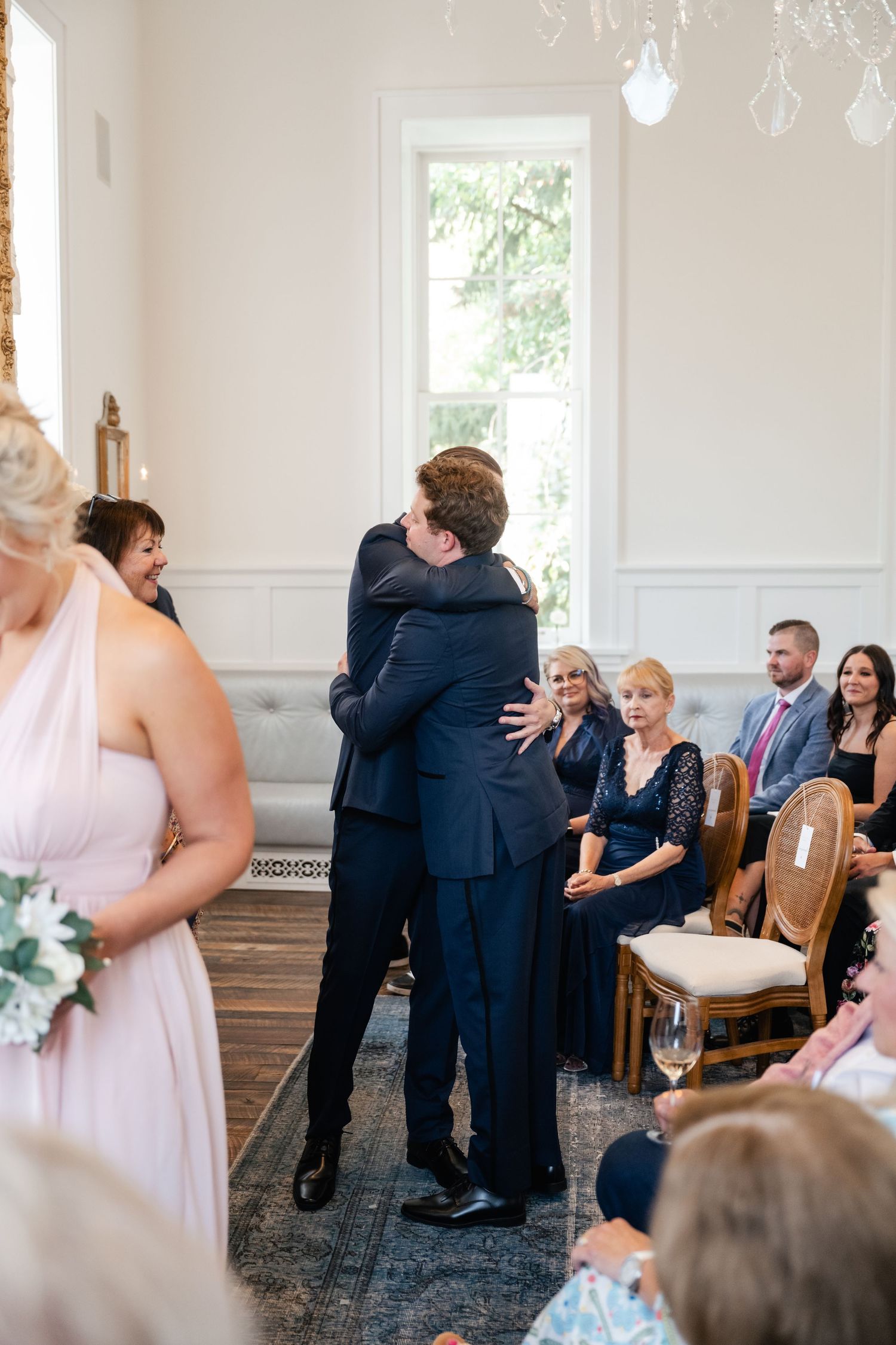 Emotional moment during wedding ceremony with guests seated in white room with tall windows and wooden floors.