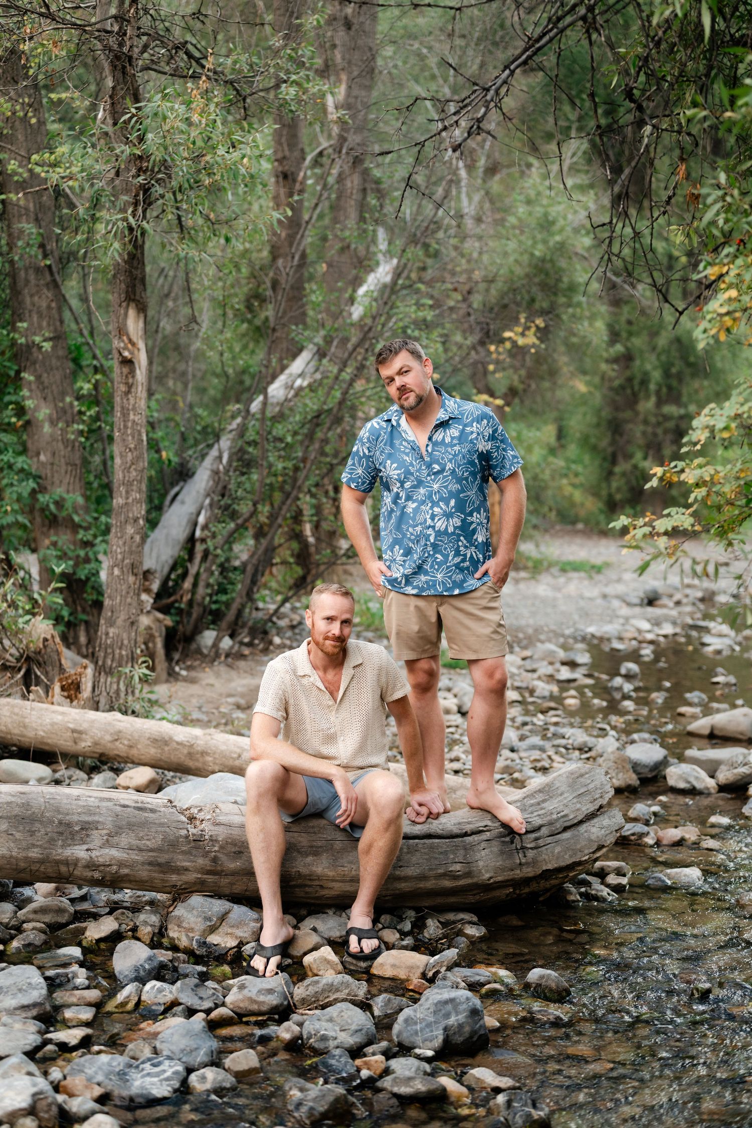 Two men pose on a fallen log by a rocky creek in a lush forest, one sitting and one standing. Gay couple engagement photos in American Fork.