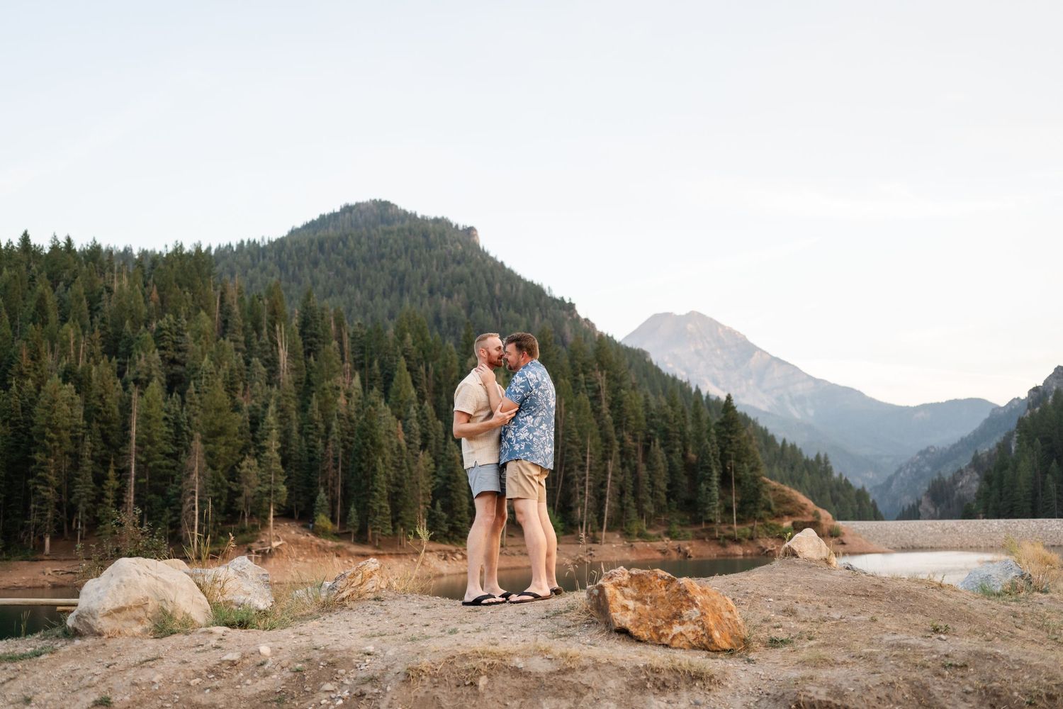 Gay engagements at Tibble Fork. Couple share a romantic moment on a rocky overlook with a mountain lake and evergreen forest backdrop.