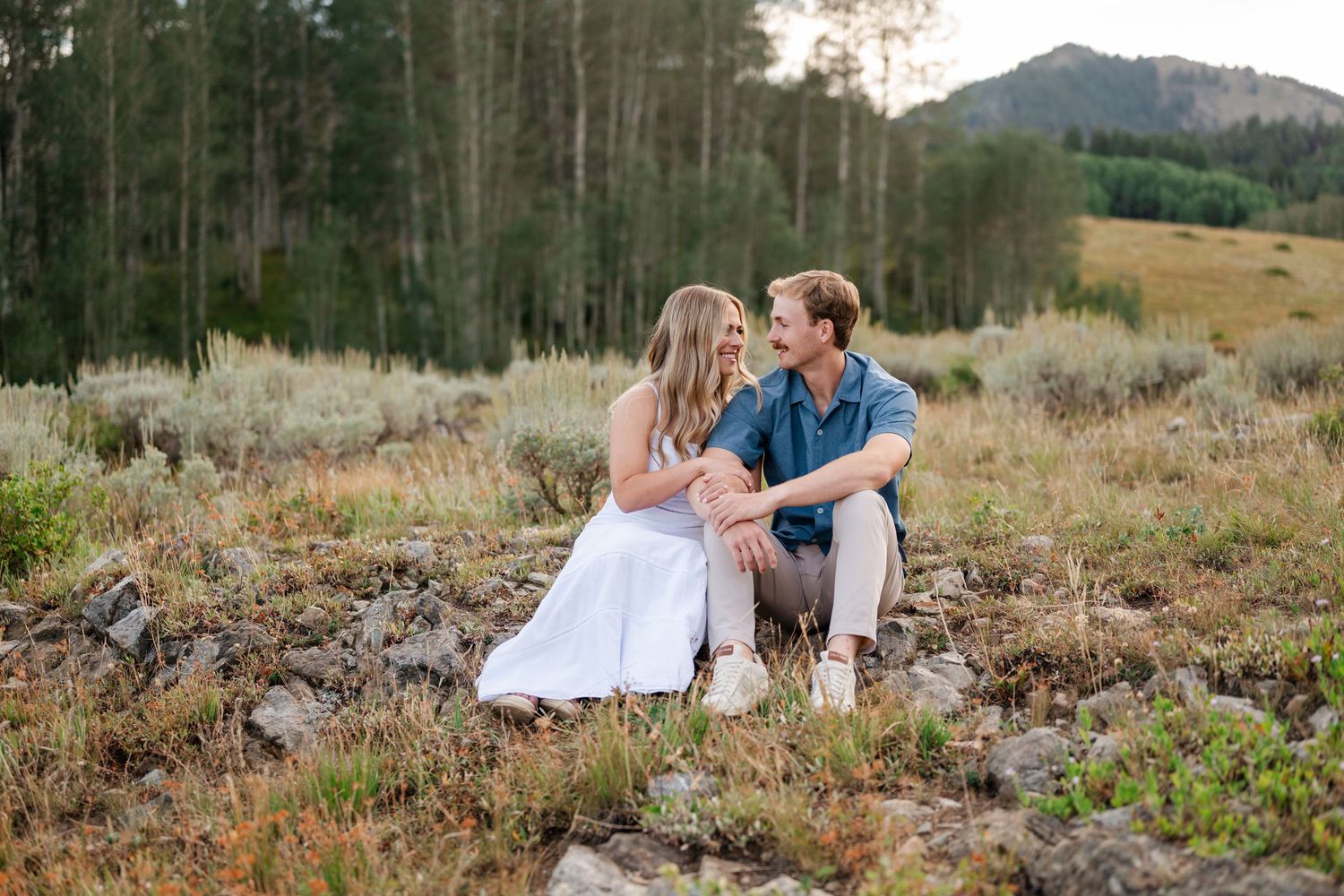 Couple sitting together on rocks in a mountain meadow, smiling at each other during a golden hour engagement session.