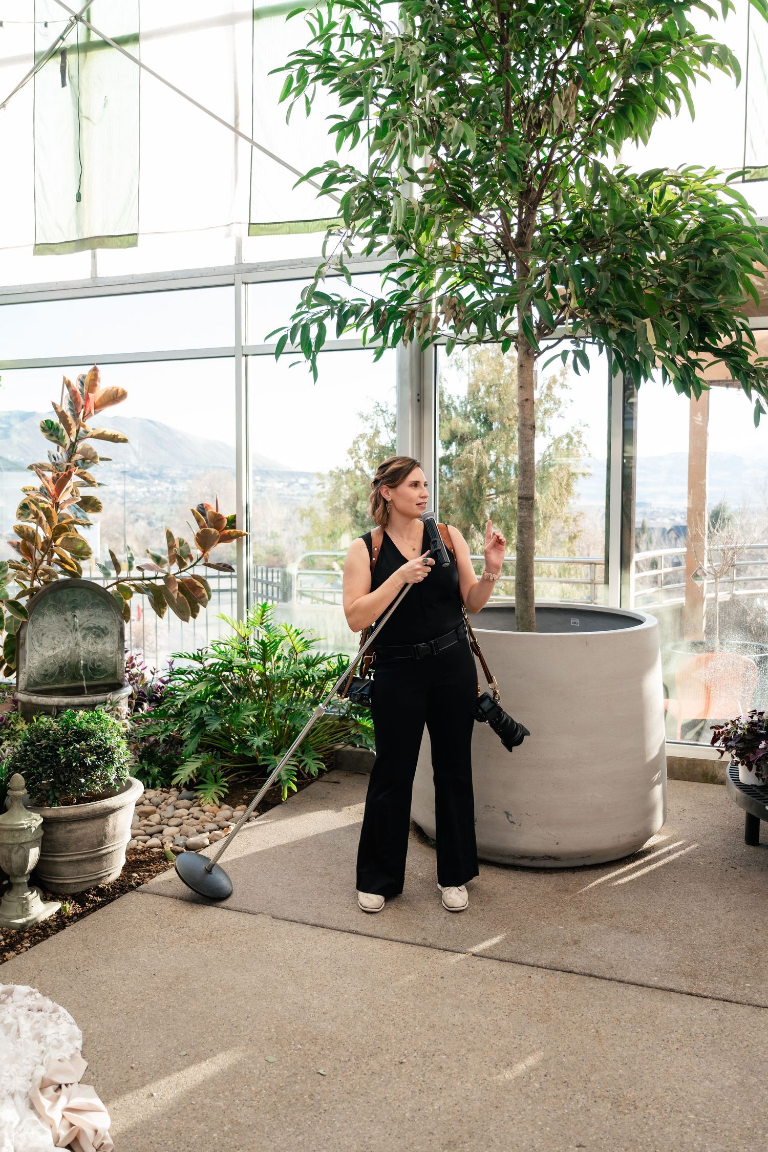 Professional photographer with camera and monopod stands among indoor plants in a bright conservatory with mountain views.