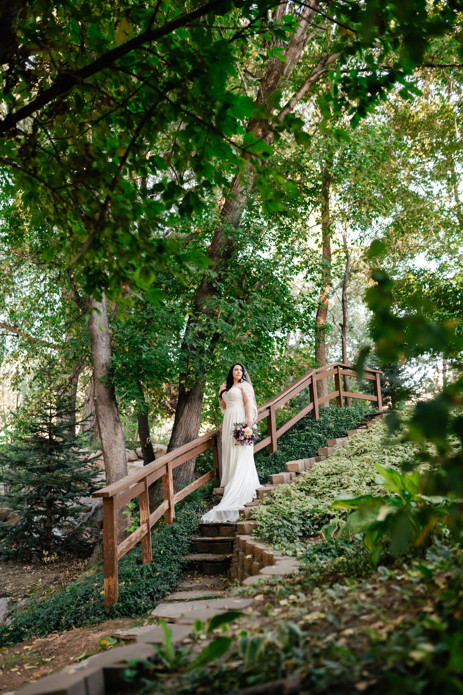 A bride in a flowing white dress stands on wooden steps surrounded by lush green trees in an outdoor forest setting.