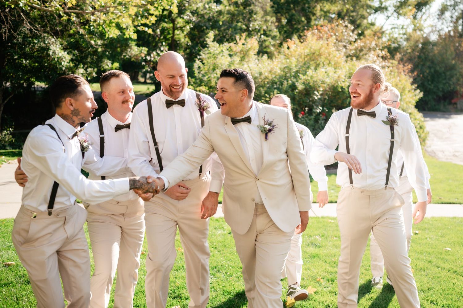 Groomsmen in light beige suits and suspenders share a candid laughing moment together on a sunny lawn.
