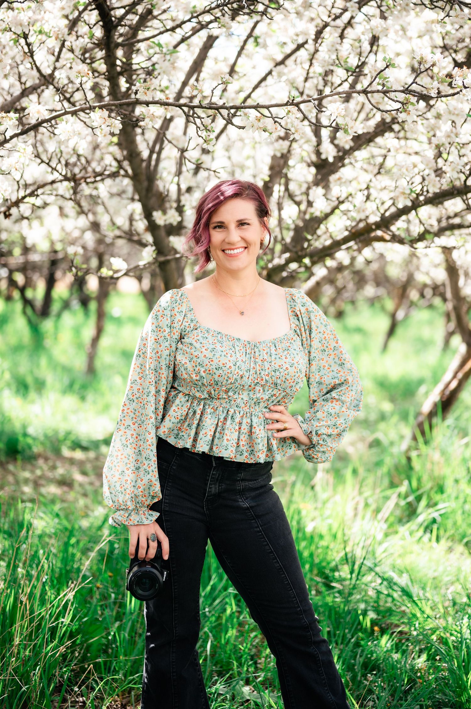 A spring portrait under blooming trees with a floral patterned top, dark jeans and pink hair against blossoms. Utah Wedding Photographer.