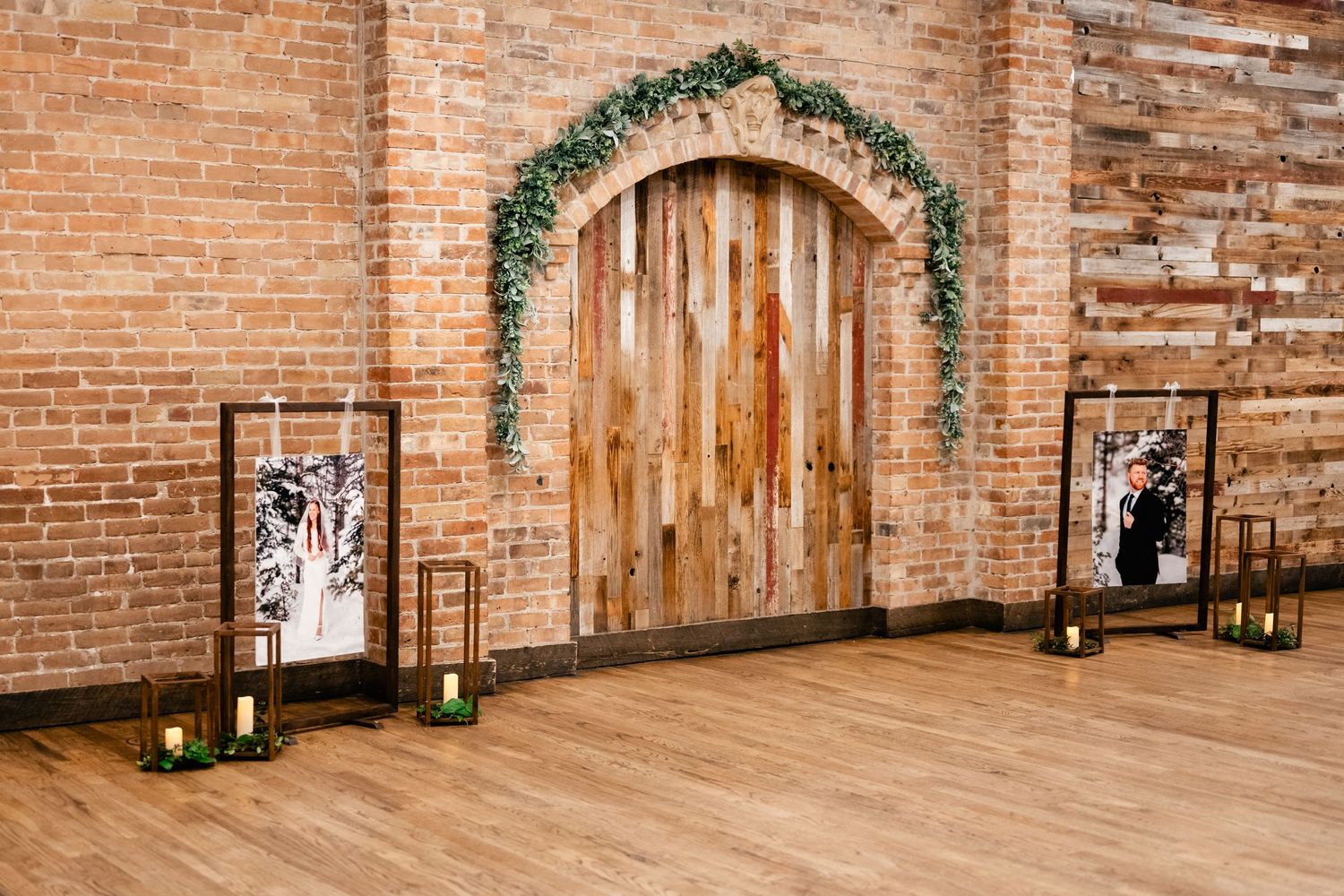 Rustic wooden door with greenery arch in exposed brick wall flanked by framed artwork and lanterns on wooden floor.
