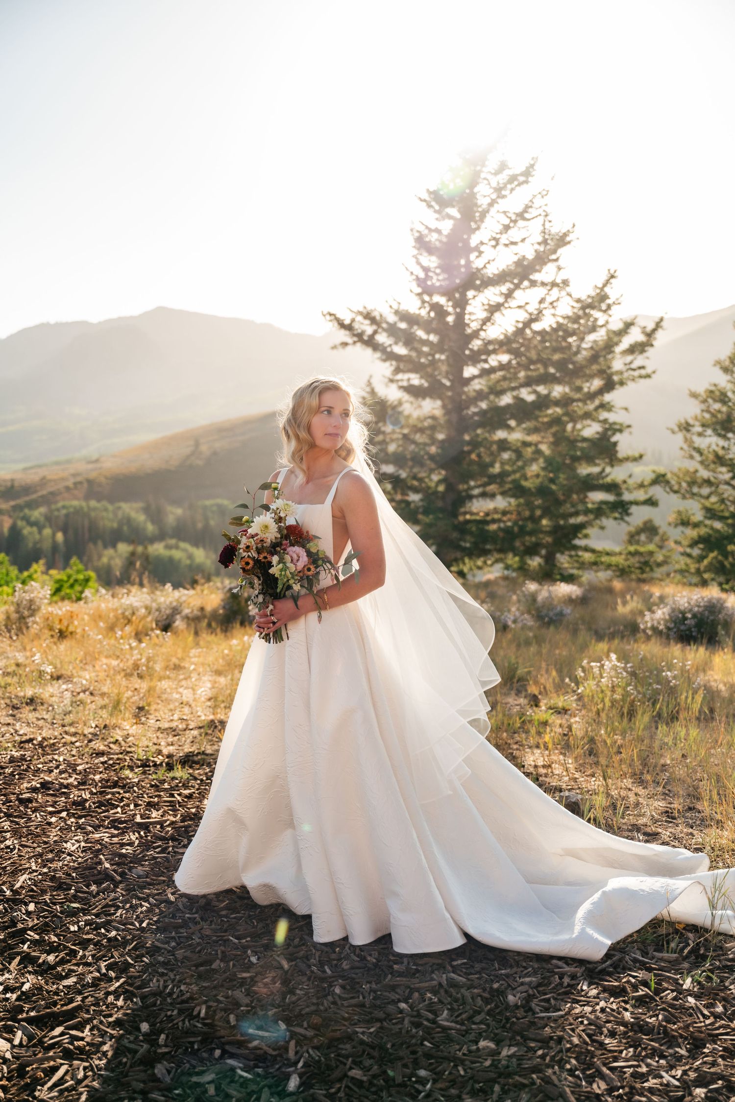 Bride in flowing white dress holds colorful bouquet against mountain landscape at golden hour. Park City Wedding Photographer. Utah Wedding.