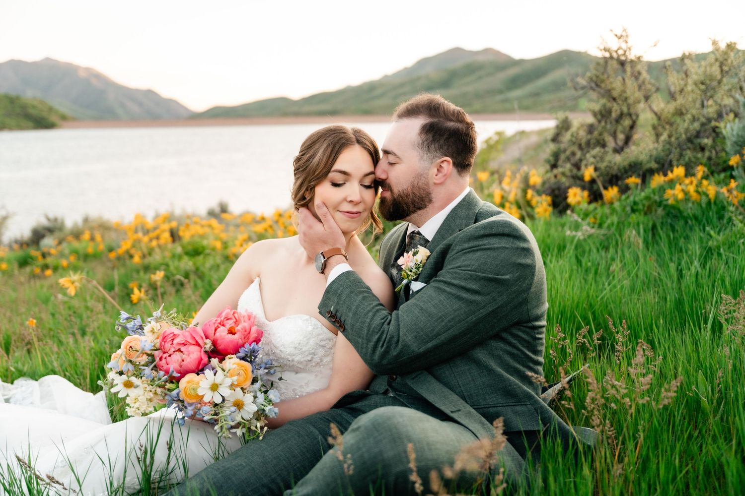 Wedding couple sits in flowering meadow holding vibrant bouquet with scenic mountain lake view.