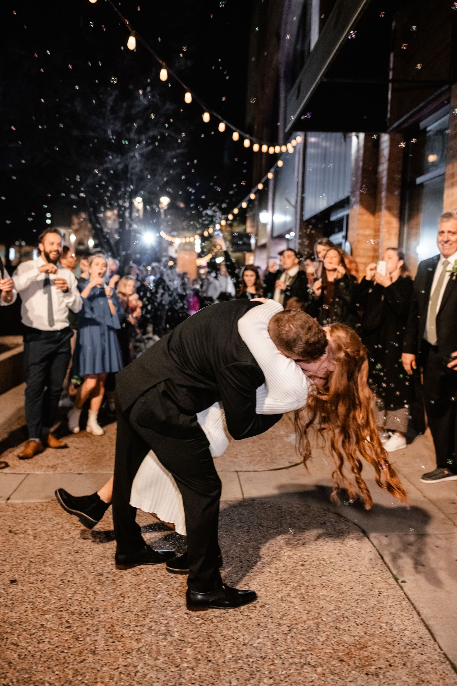 A couple shares a romantic dip kiss under string lights at an outdoor evening celebration while guests look on with sparklers.