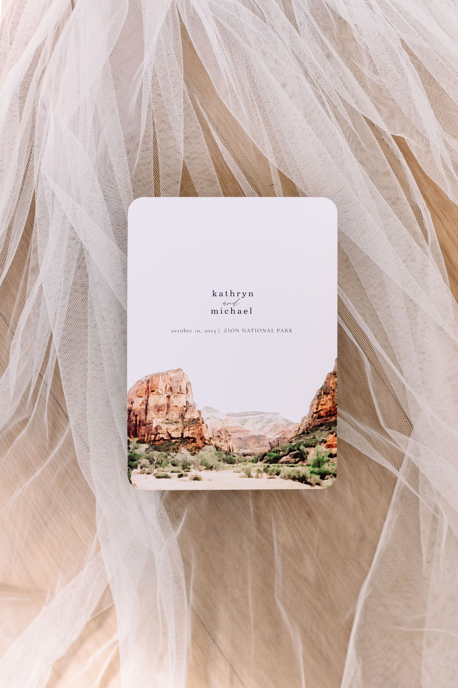 Wedding invitation card featuring desert landscape artwork against flowing white tulle fabric.