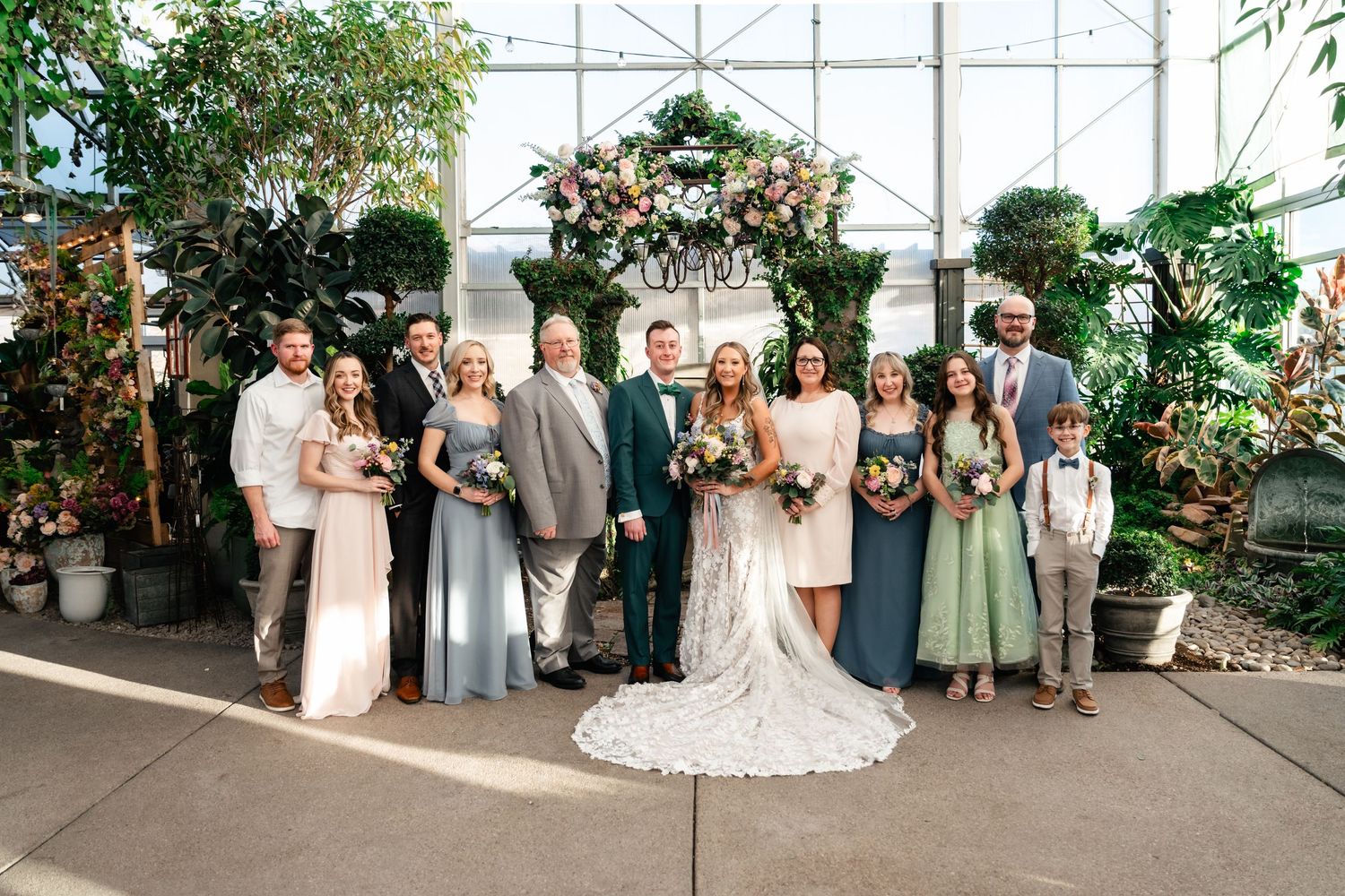 A wedding party gathers for a group photo under a floral arch in a greenhouse with decorative potted plants.