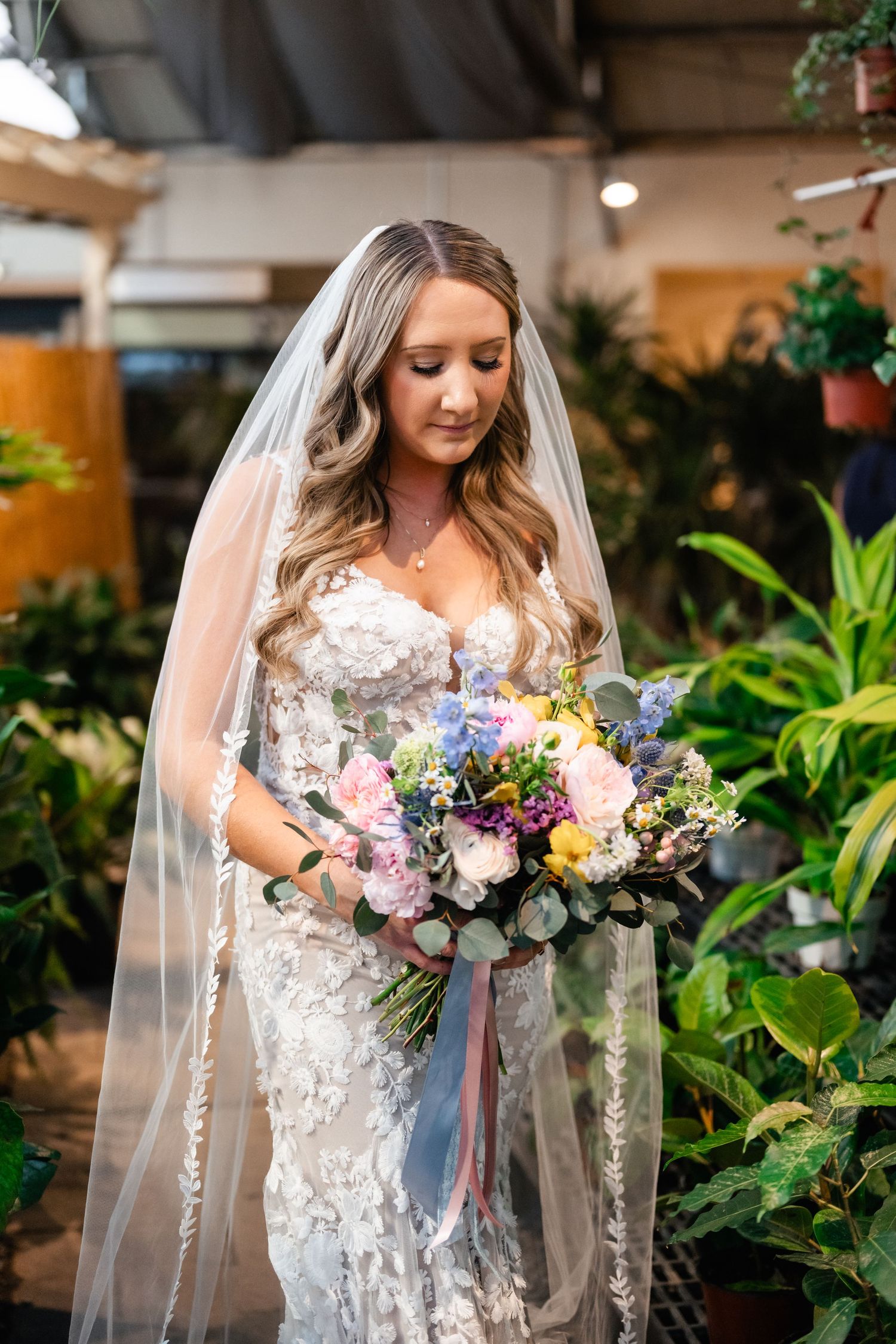 Bride in lace wedding dress with long veil holds beautiful pastel bouquet of mixed flowers in indoor garden setting.