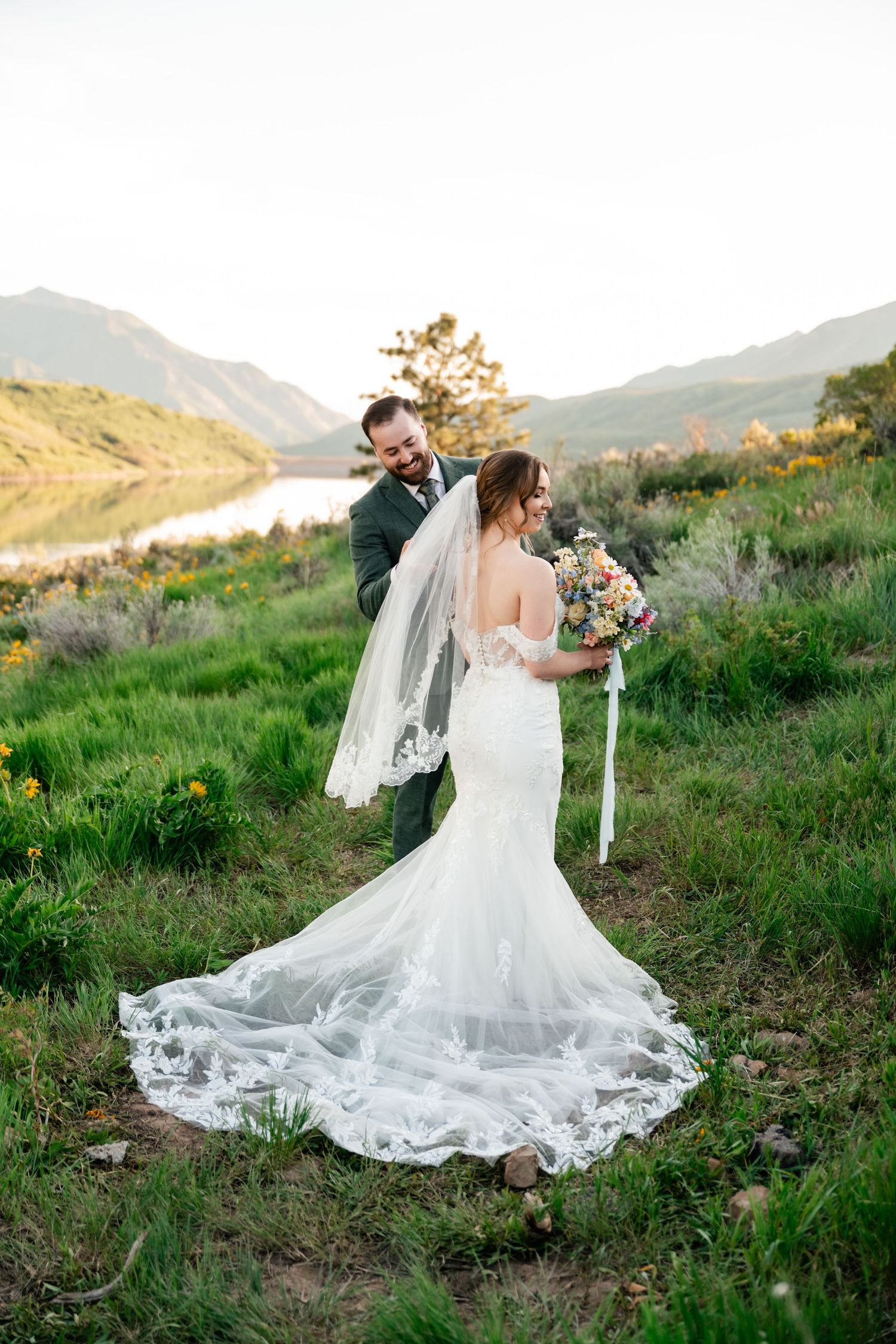 Couple shares intimate moment on grassy hillside overlooking lake with flowery bouquet and long lace wedding train.