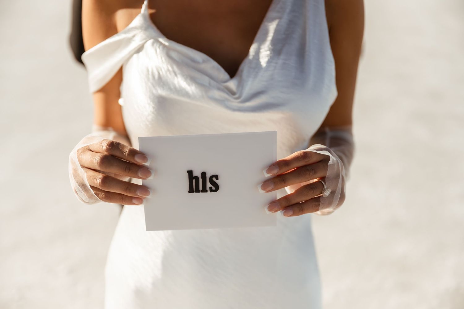 Person in white dress holding a white card with 'his' text against a light background.