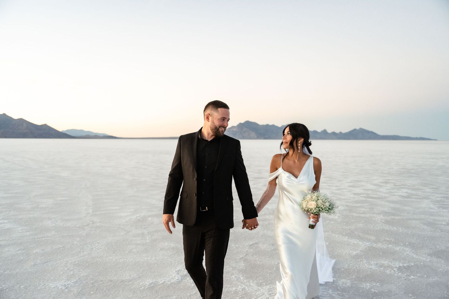 A couple walks hand in hand across the white salt flats during their wedding photoshoot at sunset.