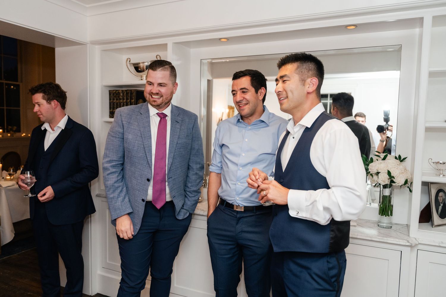 Wedding guests in formal attire socialize in an elegant white-walled room.