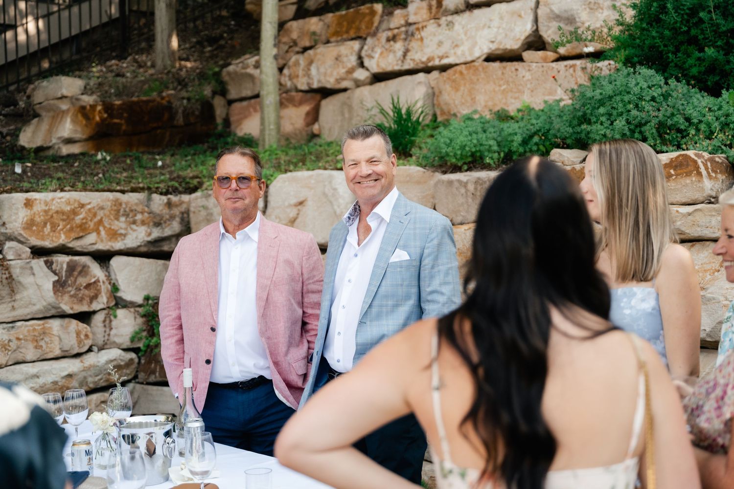A group of wedding guests sit at an outdoor reception table near a rustic stone wall.
