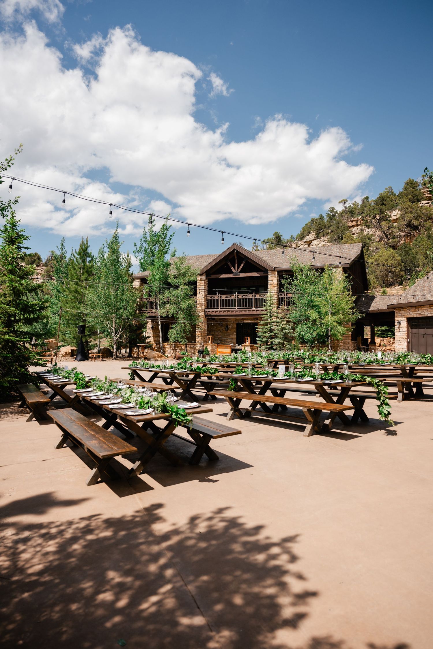 Rustic outdoor wedding venue with wooden picnic tables arranged under string lights and mountain views.