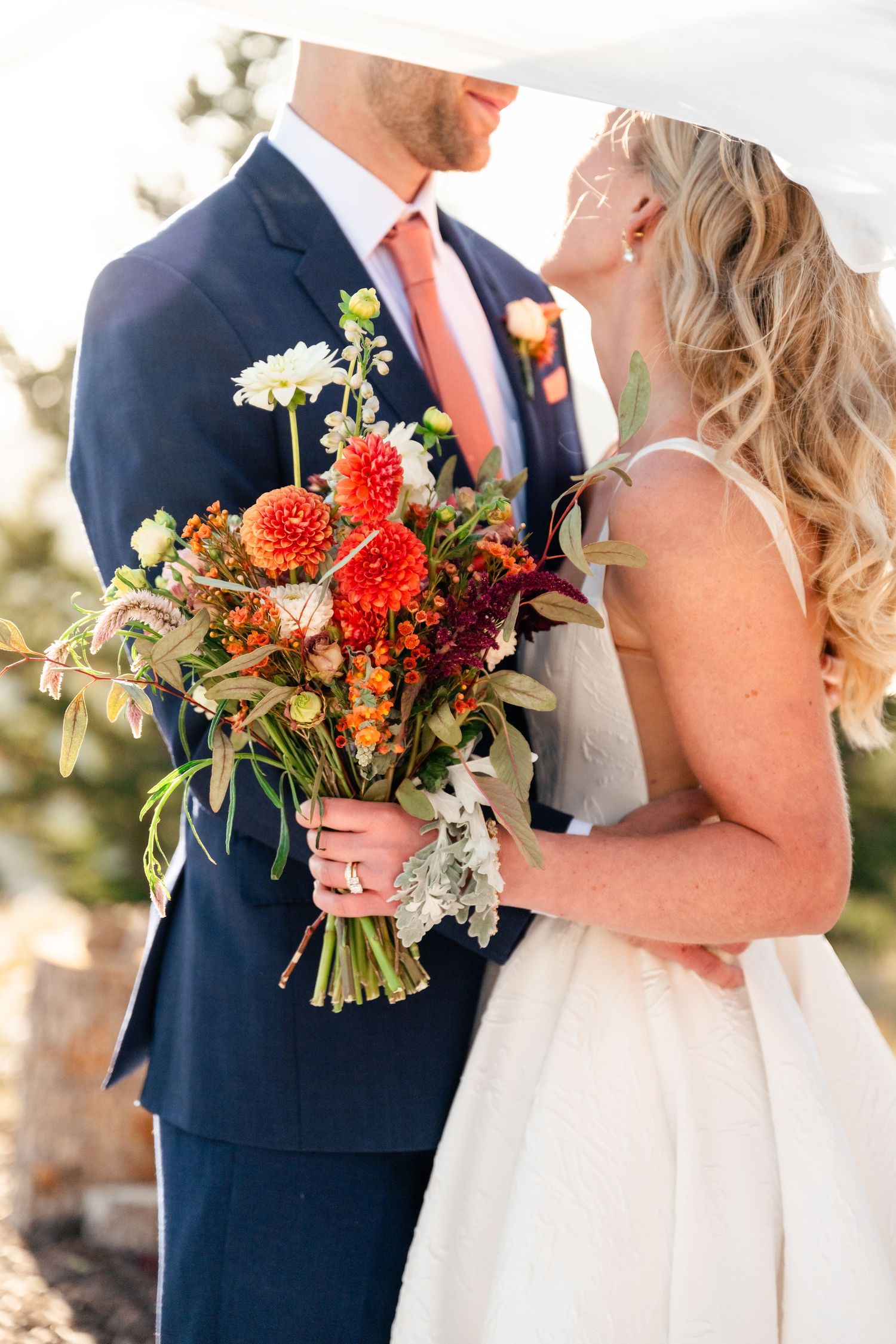 Wedding couple embraces outdoors with vibrant orange and red fall bouquet against natural scenery.