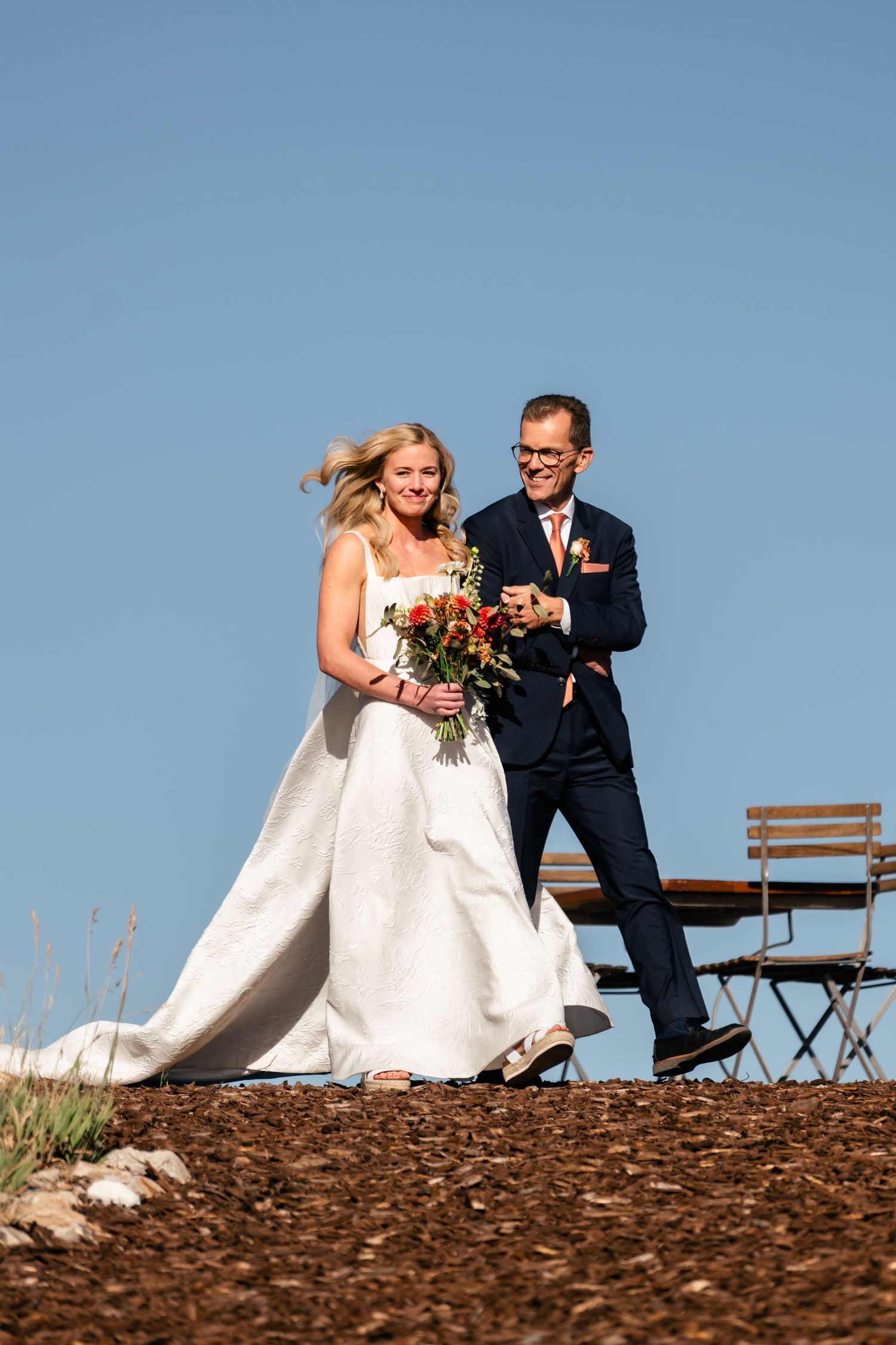 Wedding couple walking together on a hilltop against blue sky with flowing white dress.
