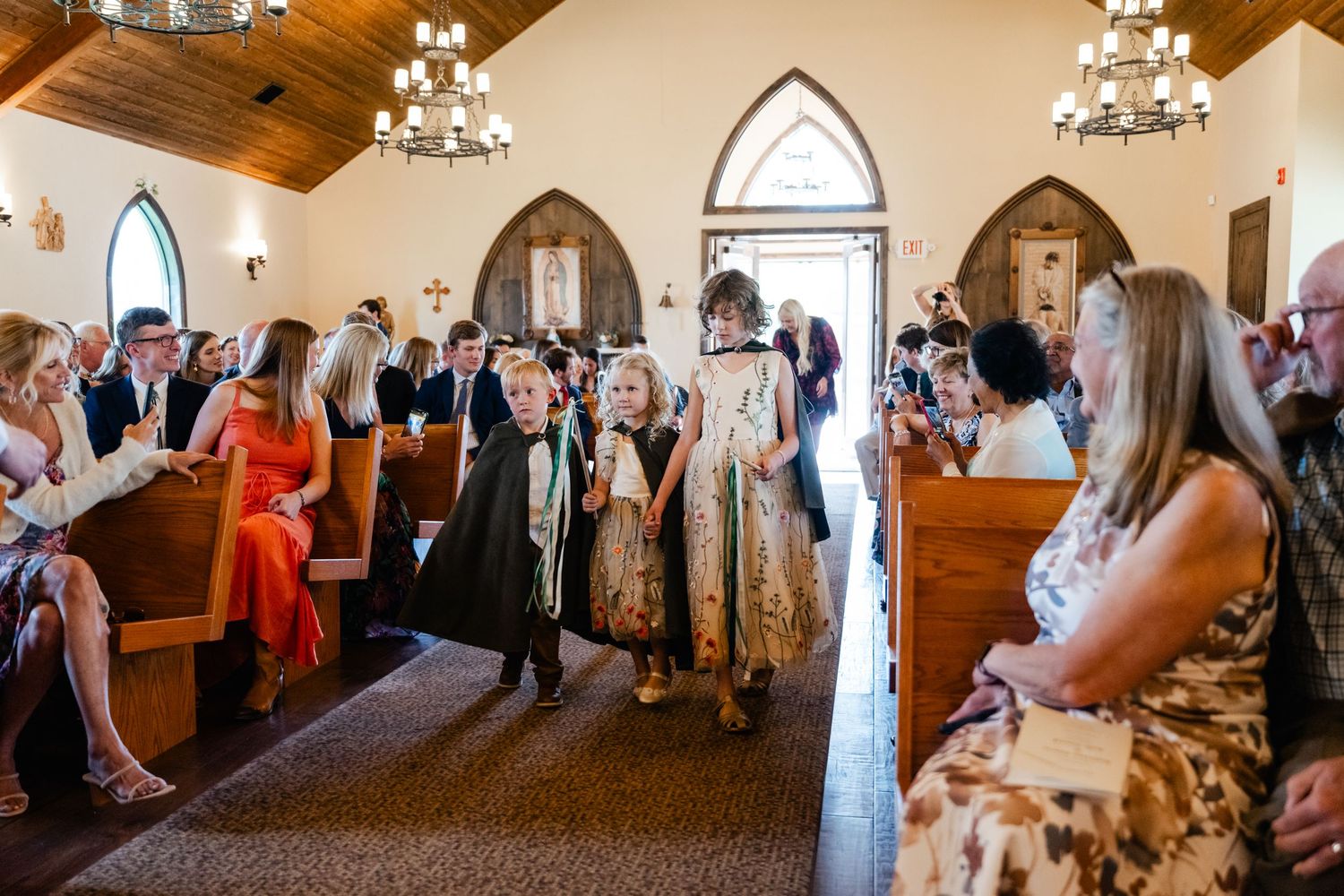 Wedding guests stand in church pews during a ceremony in a beautiful chapel with wooden beams and arched windows.